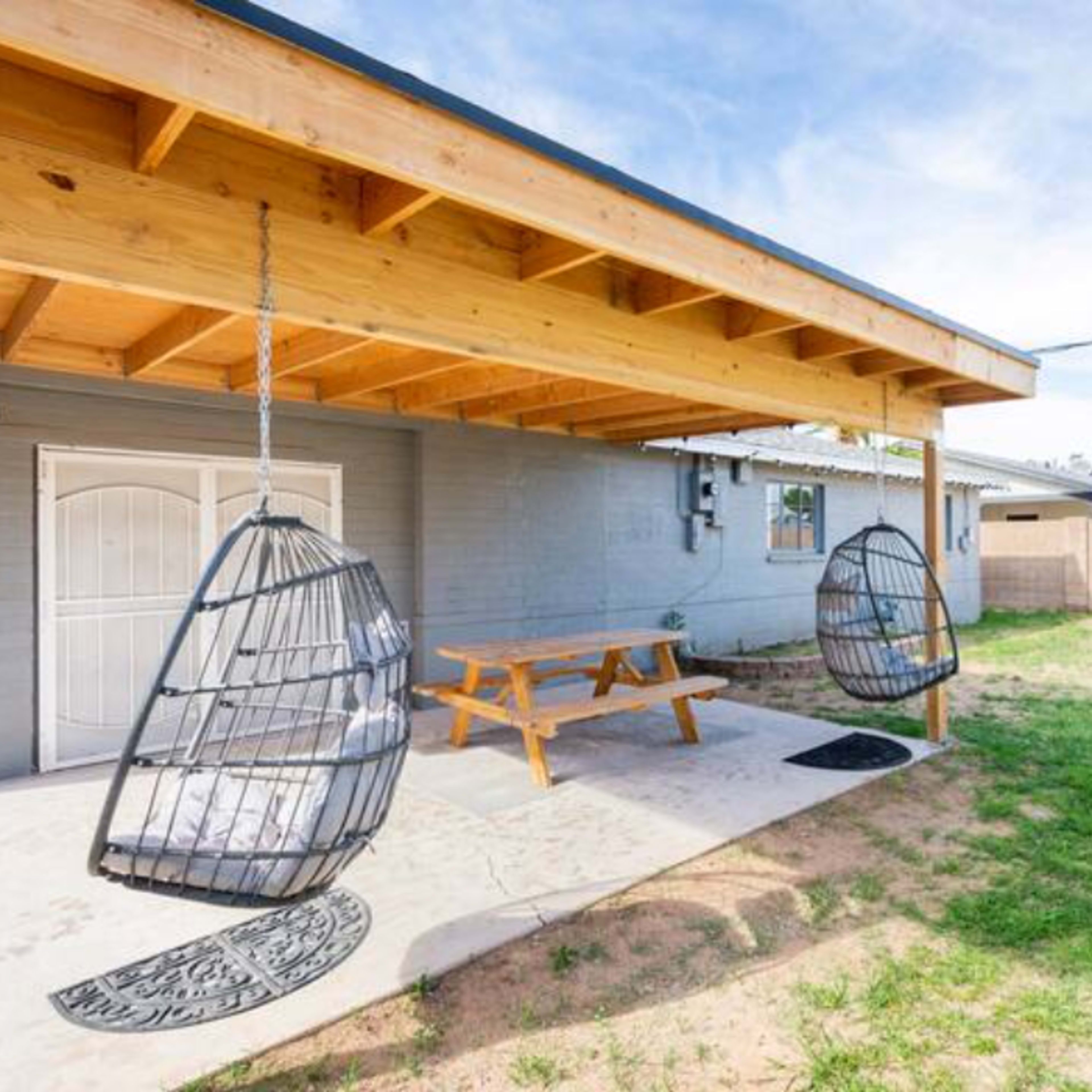 A backyard with a covered patio featuring two hanging chairs and a wooden picnic table on a concrete surface.