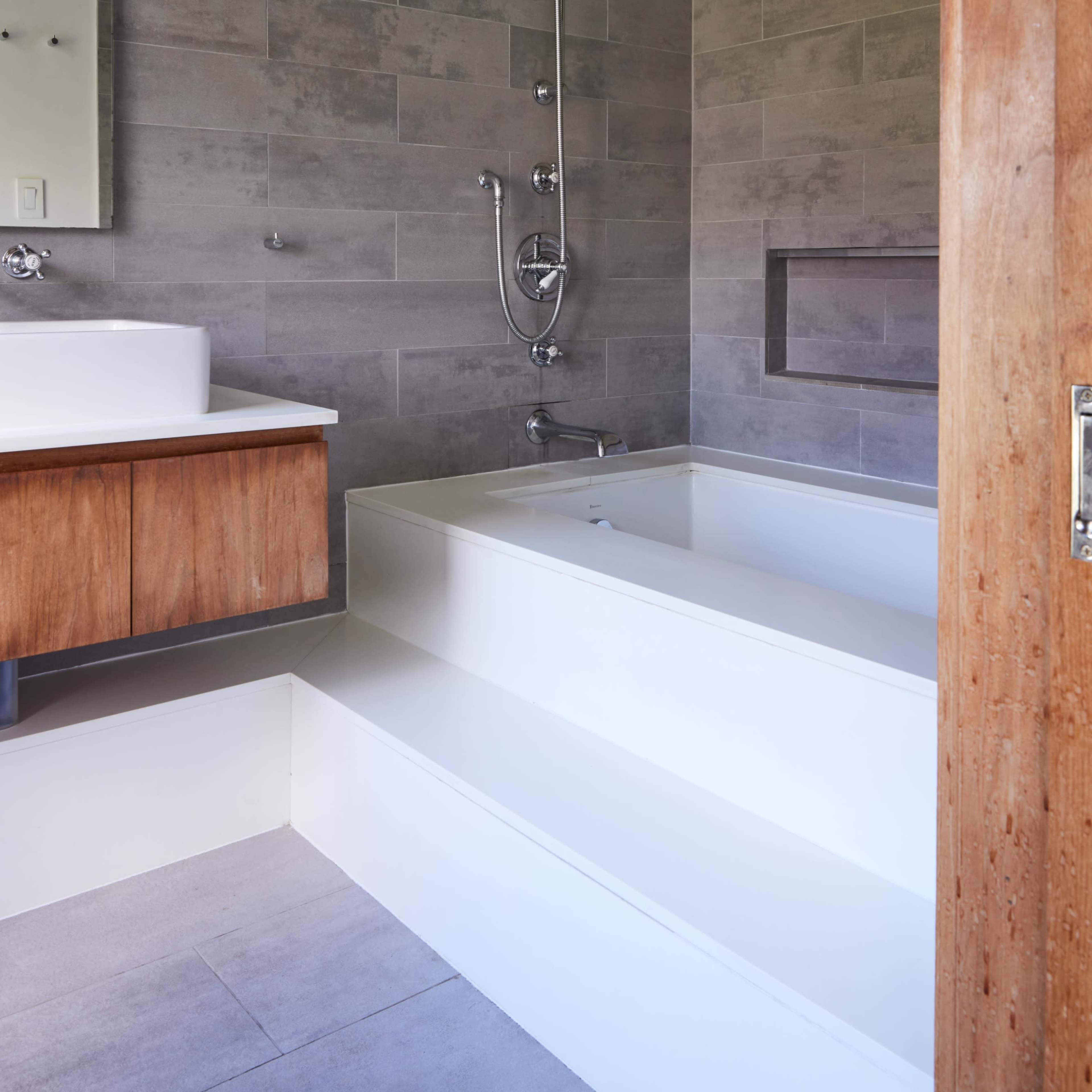 A modern bathroom featuring a shower area with a wall-mounted faucet, a rectangular sink with a wooden vanity, and a stepped bathtub against a gray tiled wall.