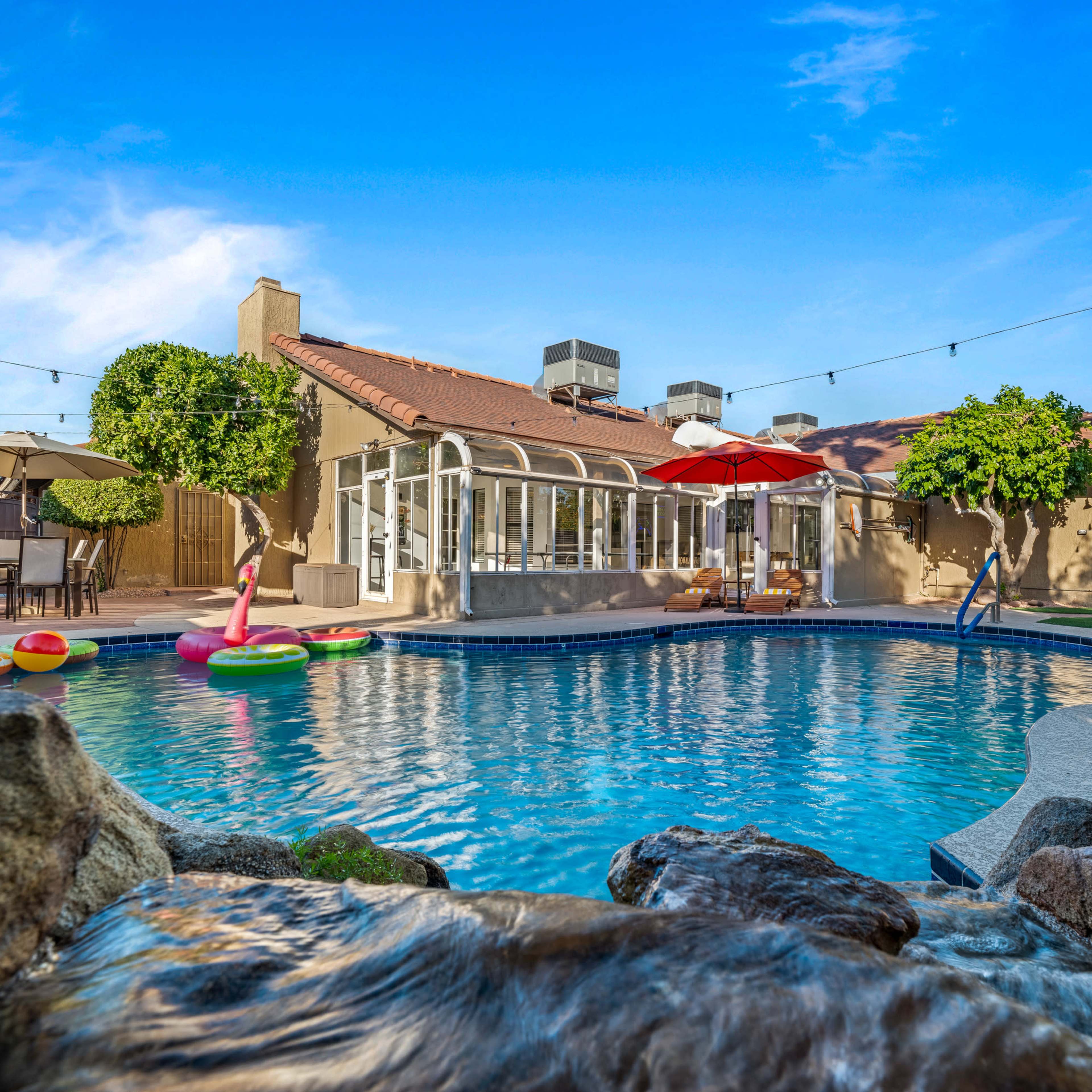 A clear pool with inflatable floats is situated in a backyard alongside a house with a patio and umbrellas.