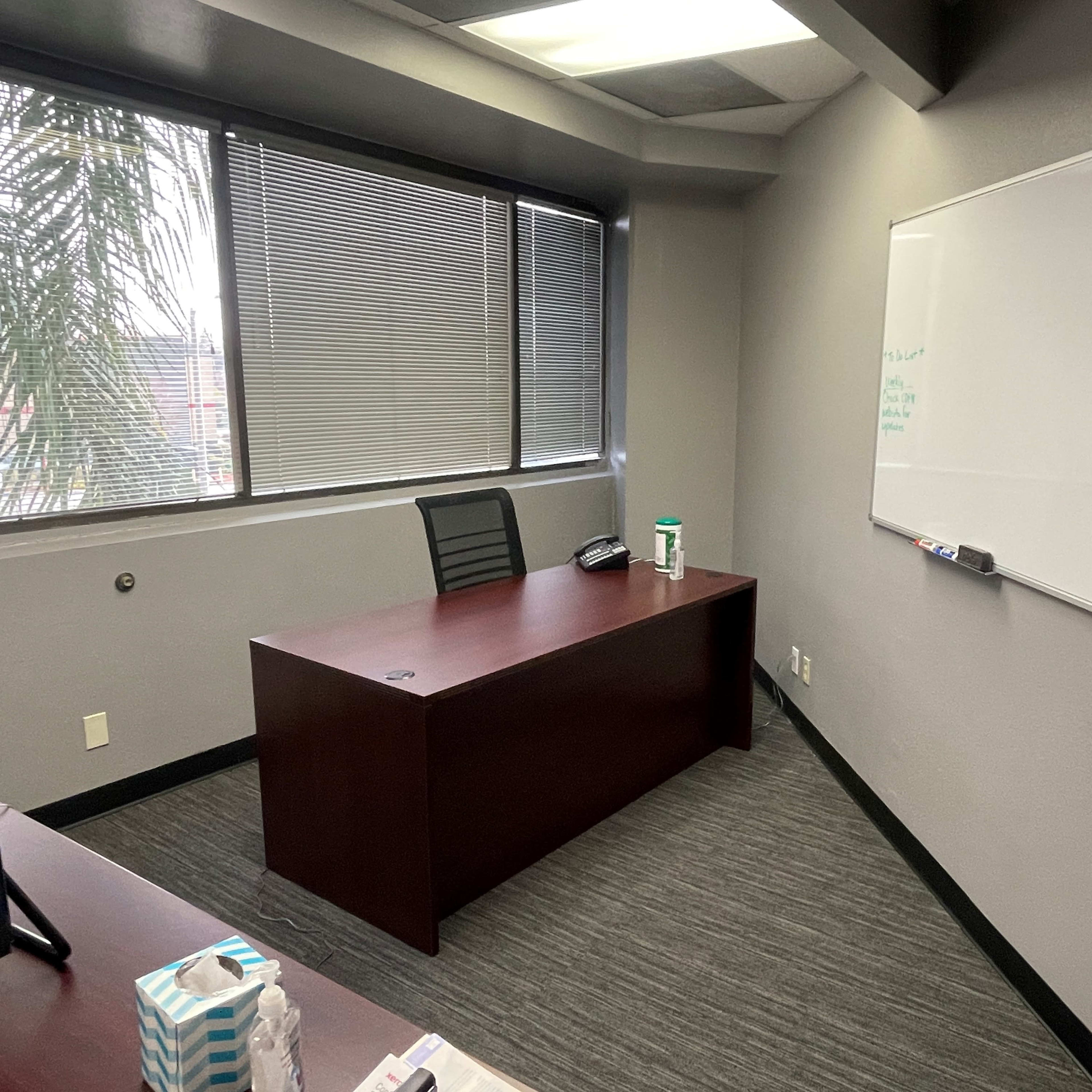A simple office space featuring a brown desk, a black office chair, a whiteboard on the wall, and a window with horizontal blinds.