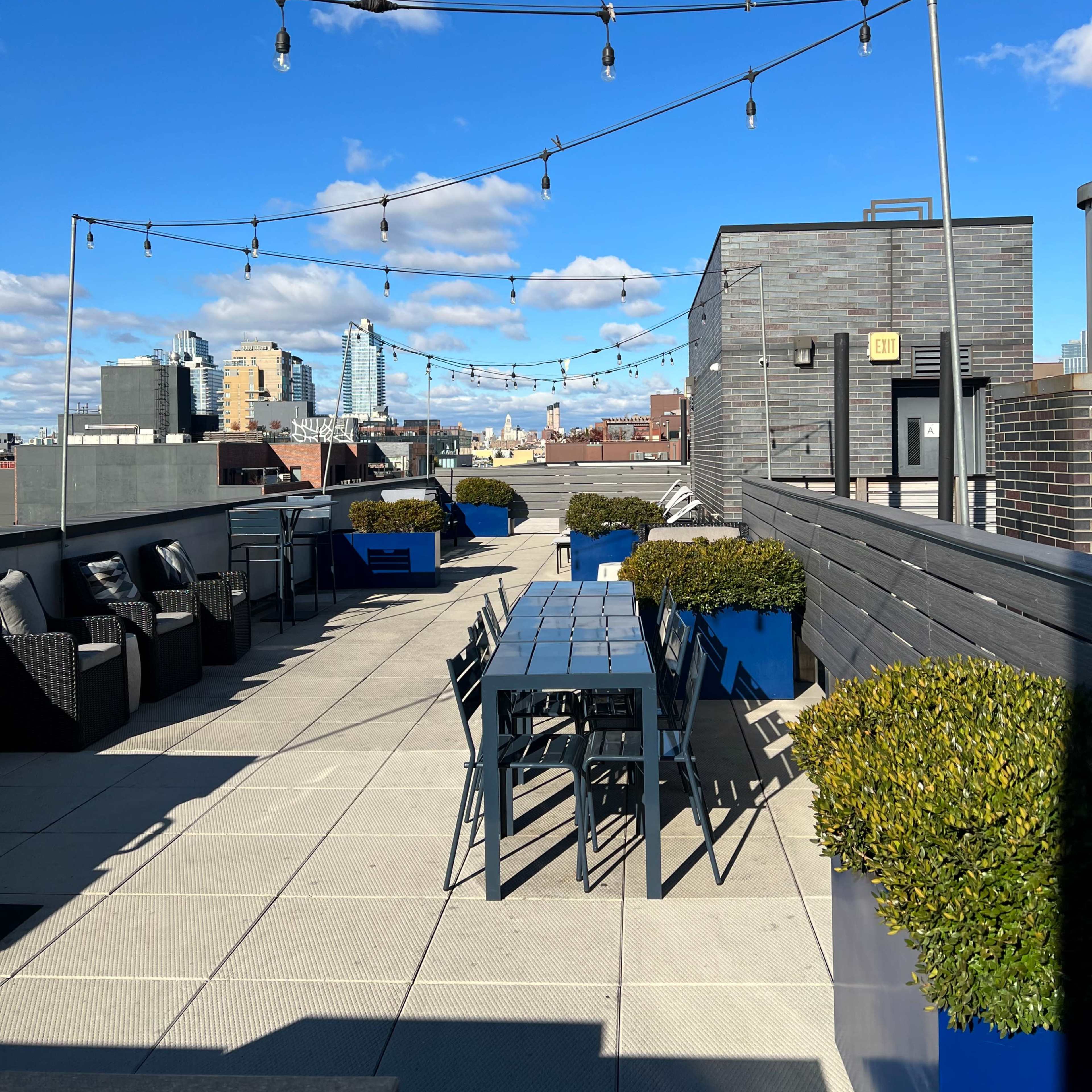 A rooftop terrace with string lights, a long dining table, and planters, set against a blue sky and city skyline.