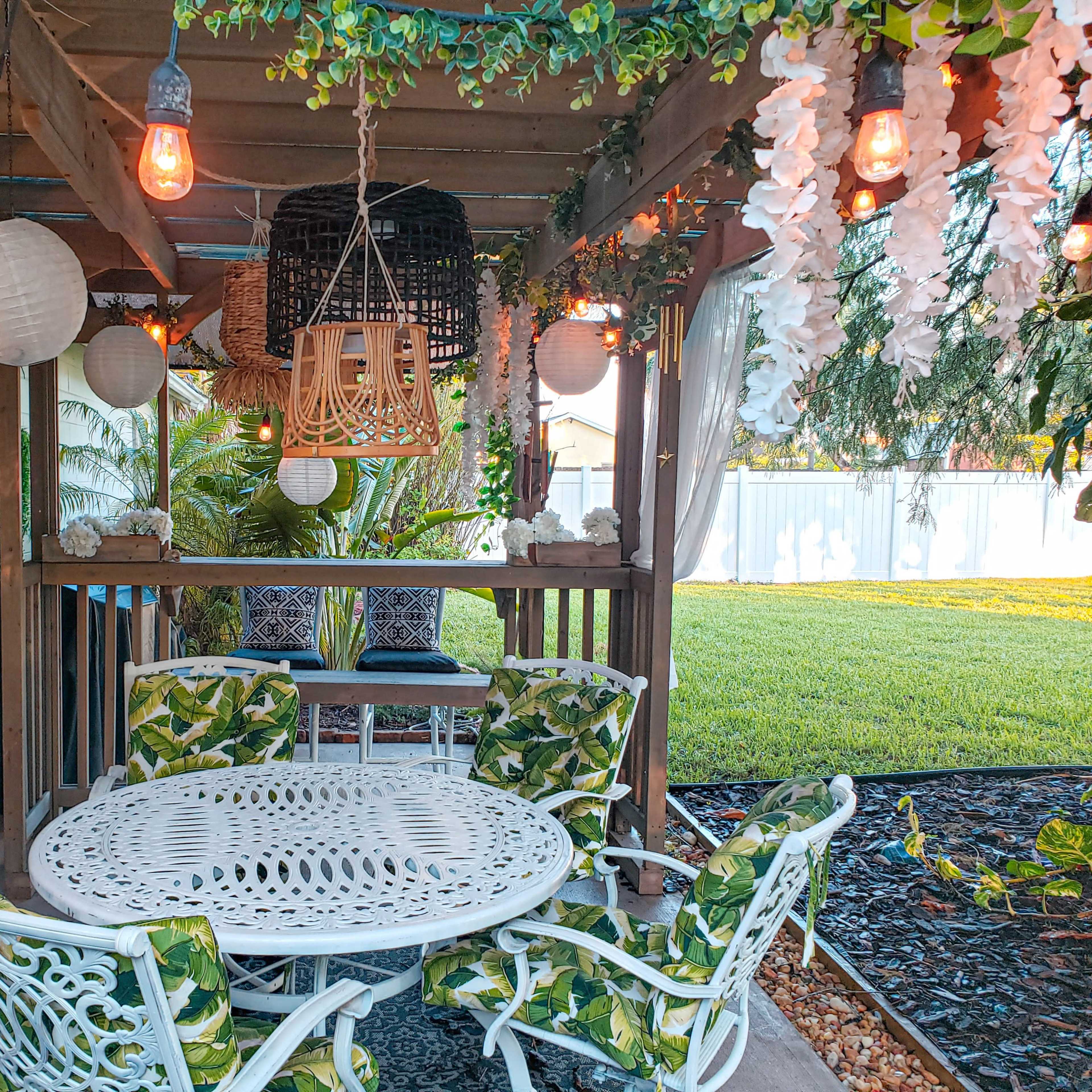 A patio with a round table and four chairs, surrounded by greenery and decorative lighting.