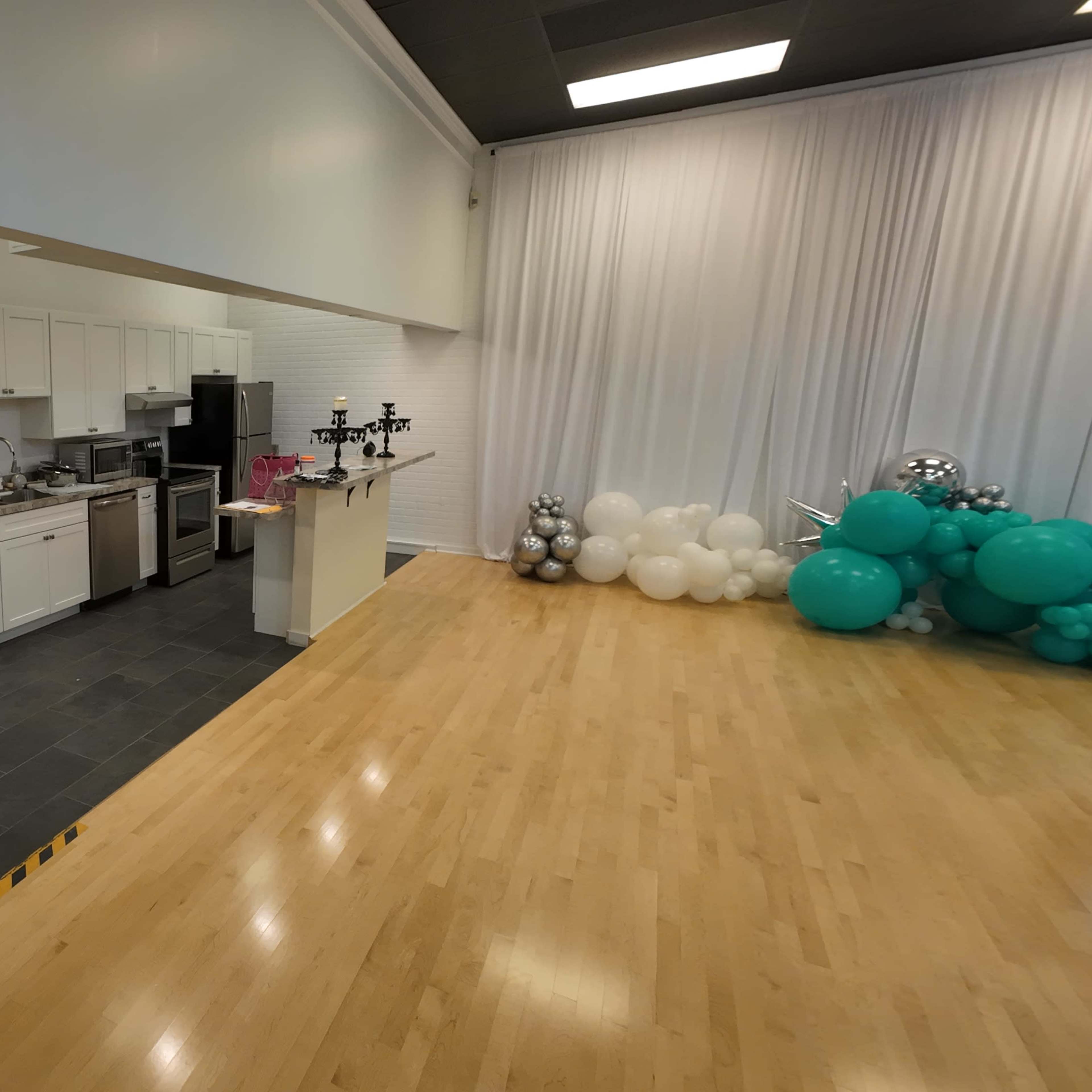 A spacious room with a kitchen area featuring white cabinets and stainless steel appliances, and a corner decorated with balloons in teal and white.