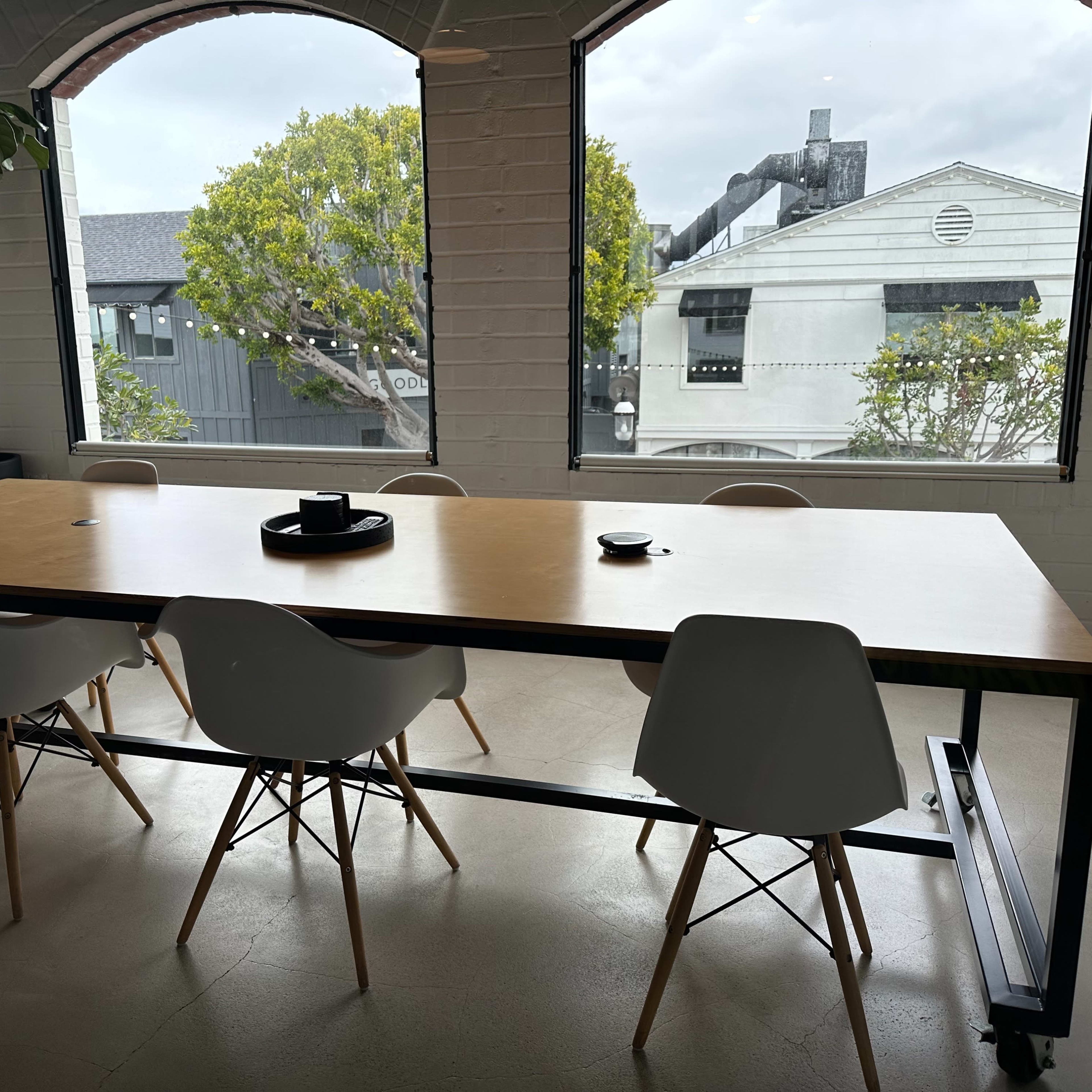 A modern meeting room features a long wooden table surrounded by white chairs, with large windows displaying a view of buildings and greenery outside.