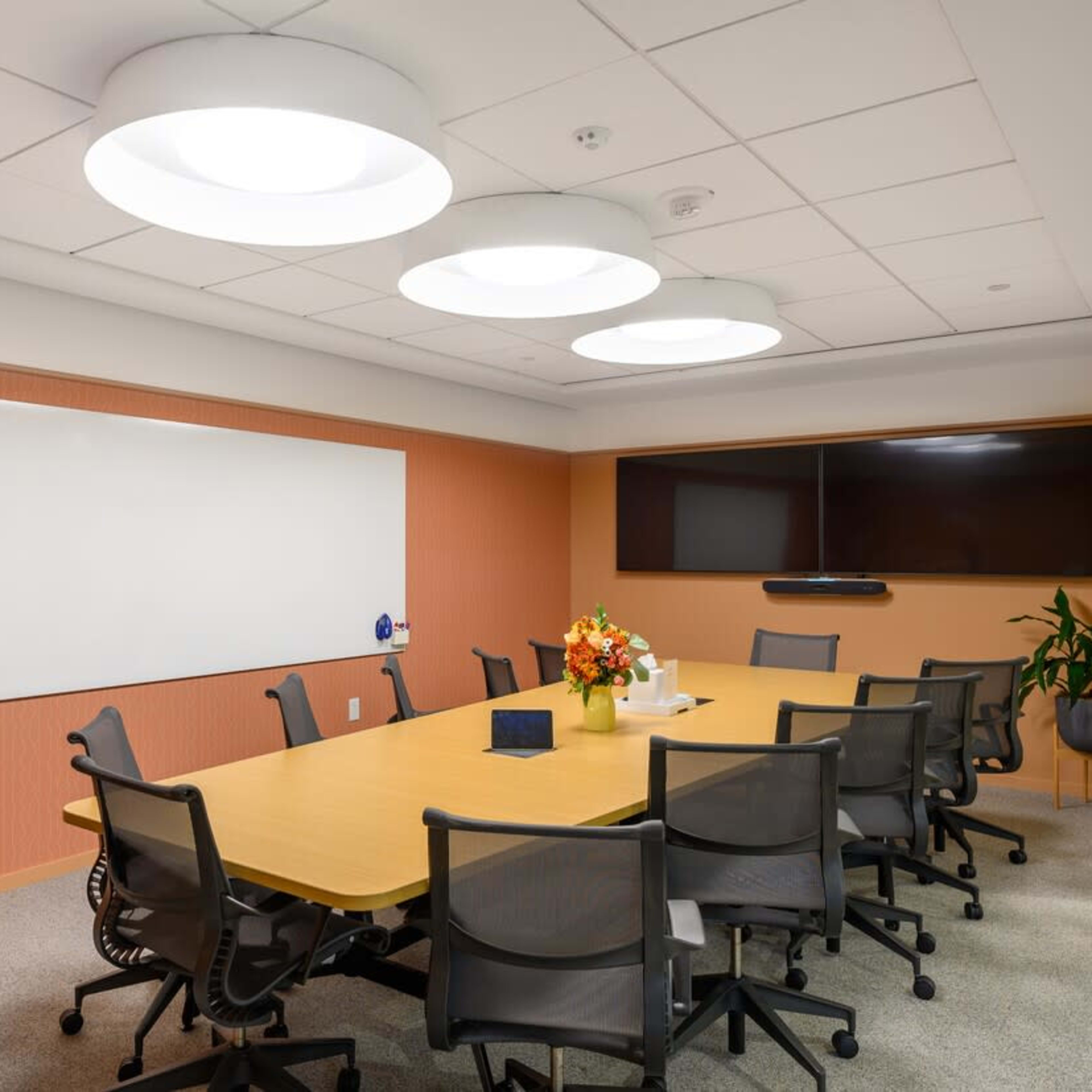 A conference room features a long wooden table surrounded by black office chairs, with a large whiteboard and a screen on the walls.
