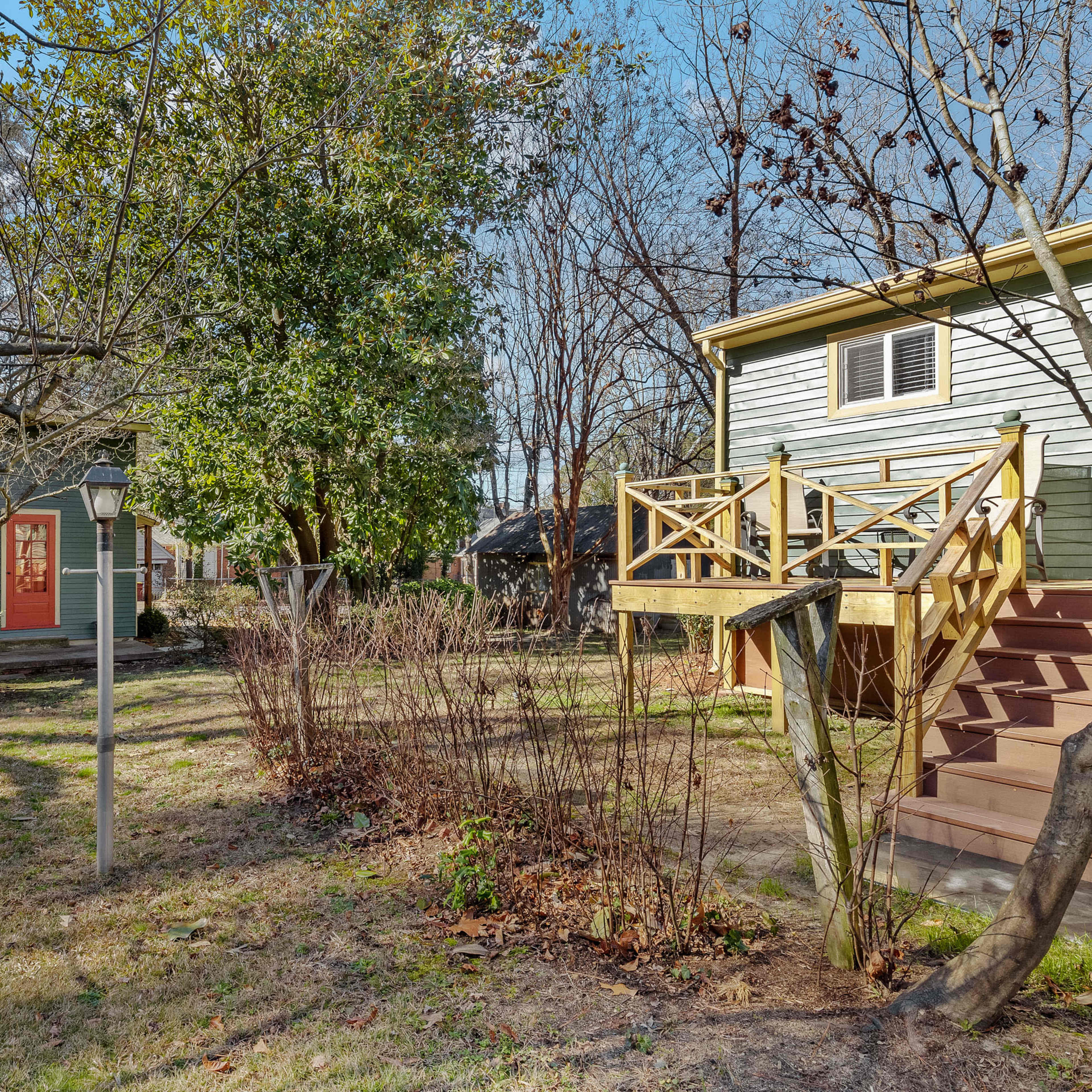 A backyard with two houses, a wooden deck with stairs, and various trees and shrubs.