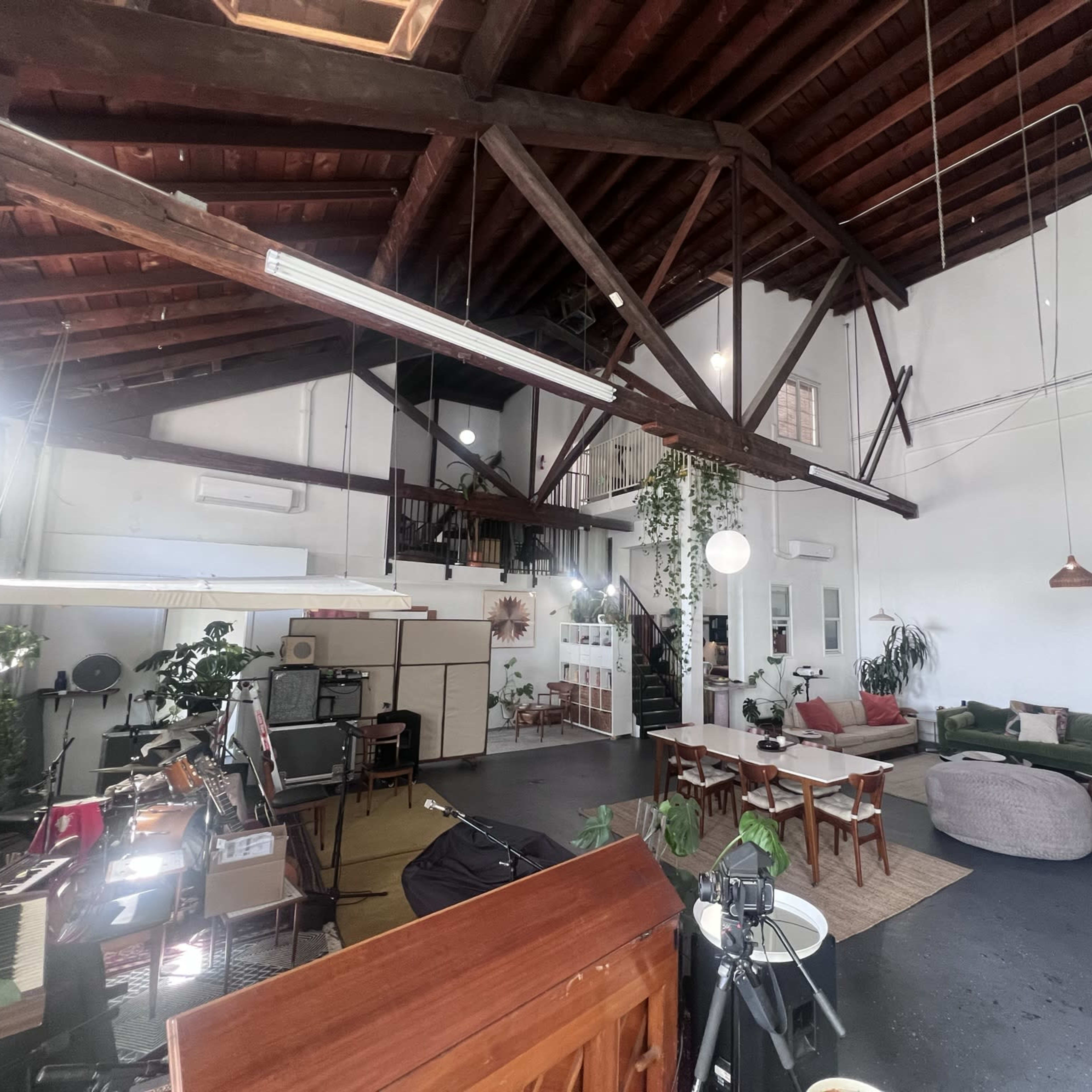 A spacious, open-concept loft with high wooden beams, a living area with a green couch, a dining space with tables, and various indoor plants throughout.