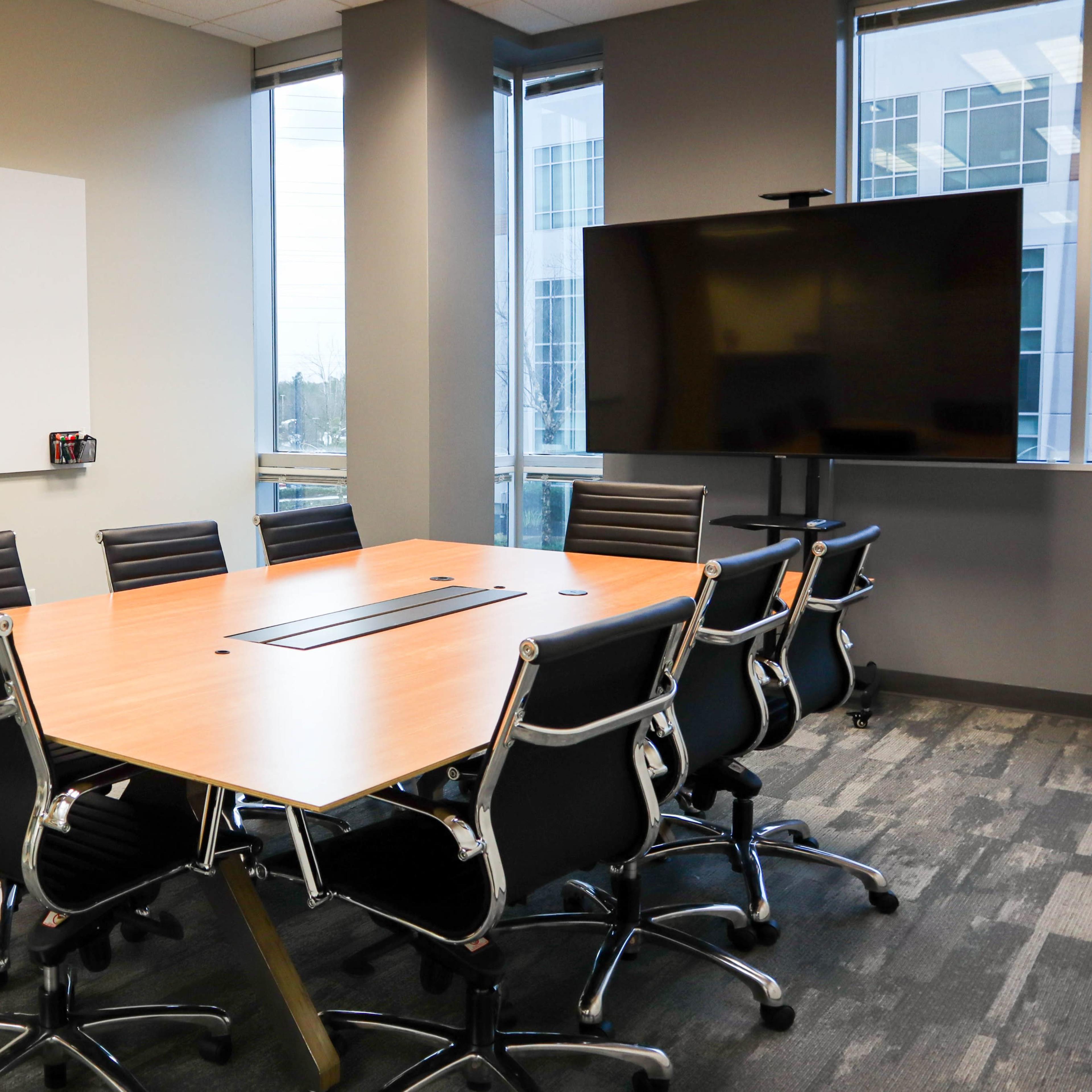 A modern conference room features a large wooden table surrounded by black office chairs, with a whiteboard and a large screen on opposite walls.