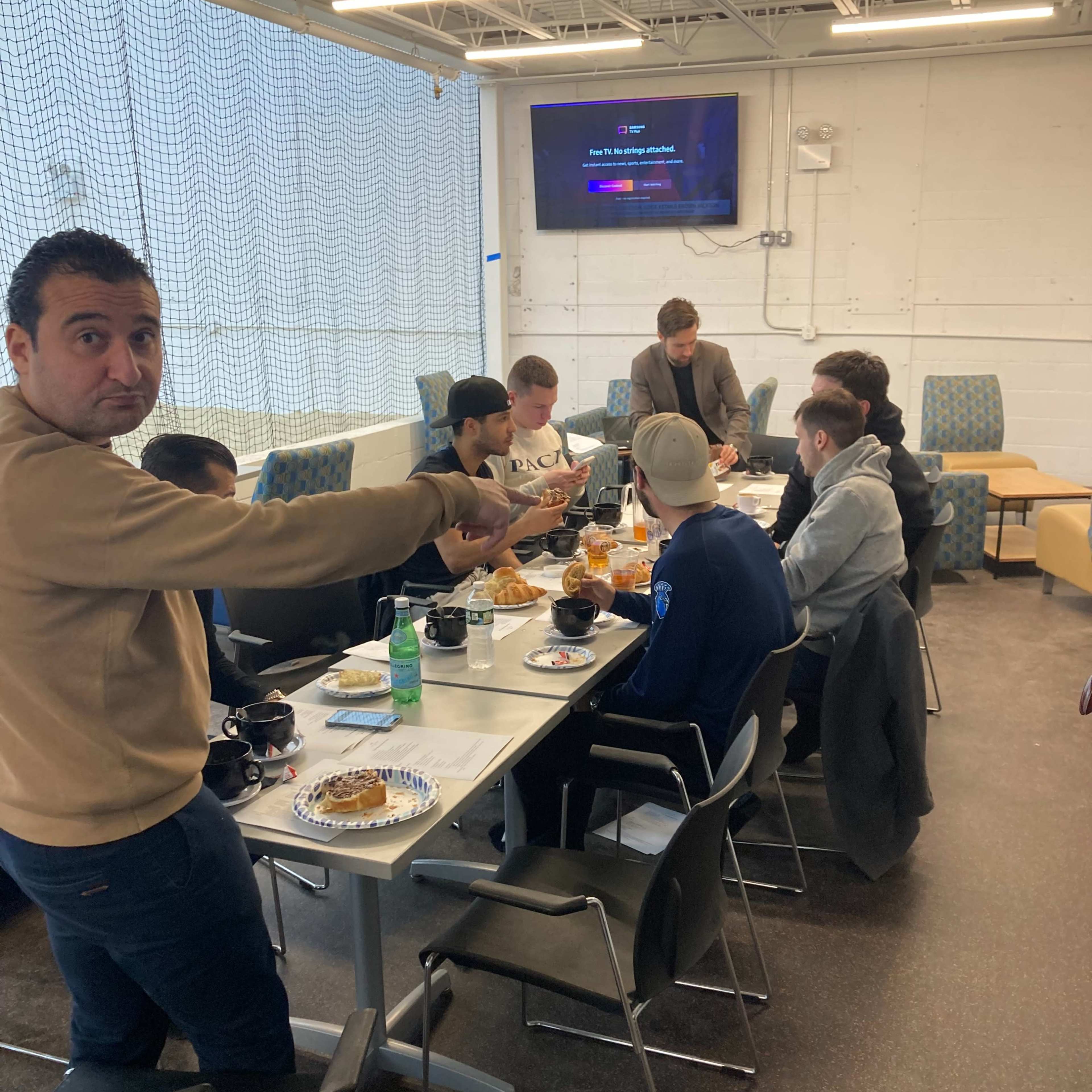 A group of people is sitting around a table filled with food, while one person is standing and gesturing towards the camera.