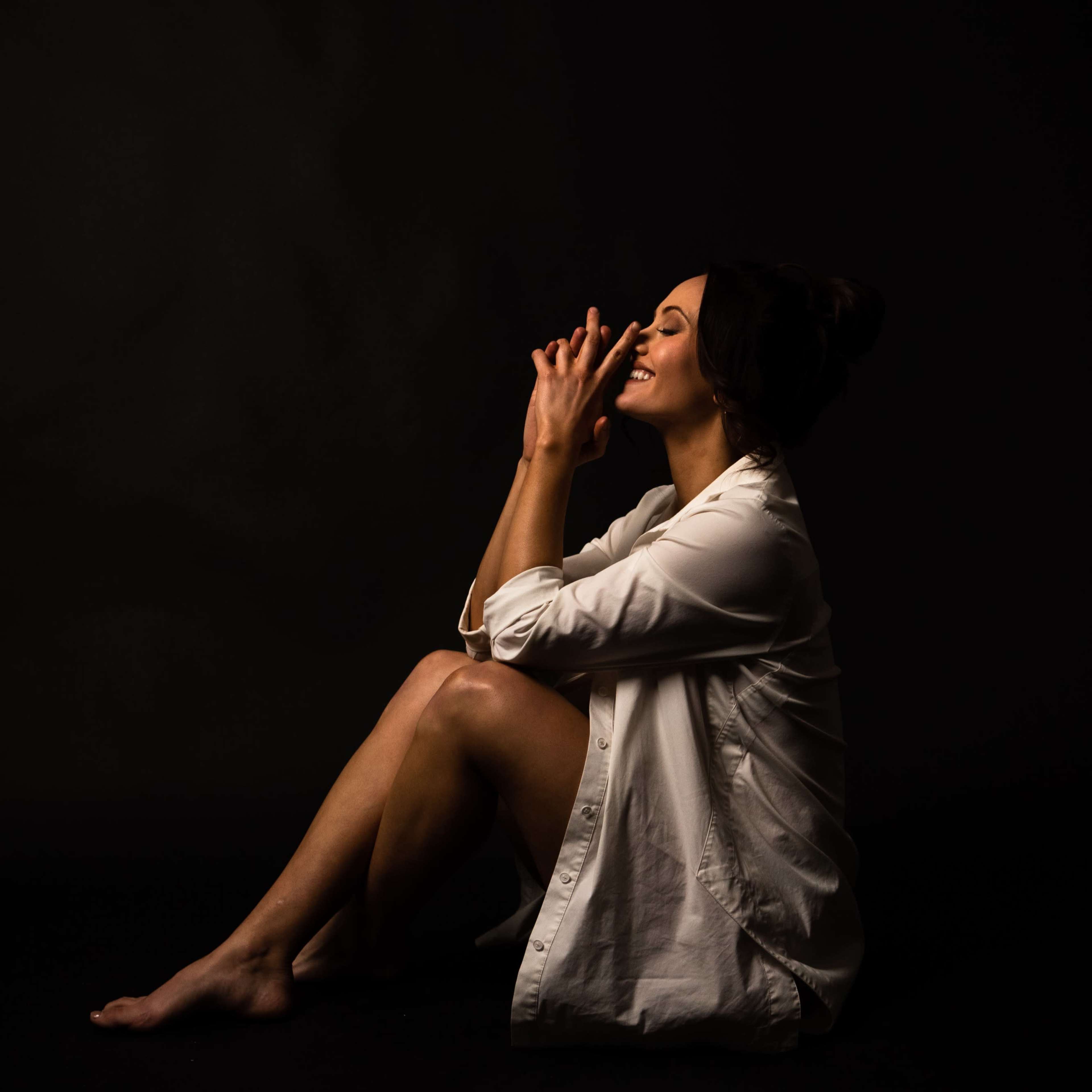 A woman in a white shirt sits on the floor, resting her chin on her hand in a dimly lit space.