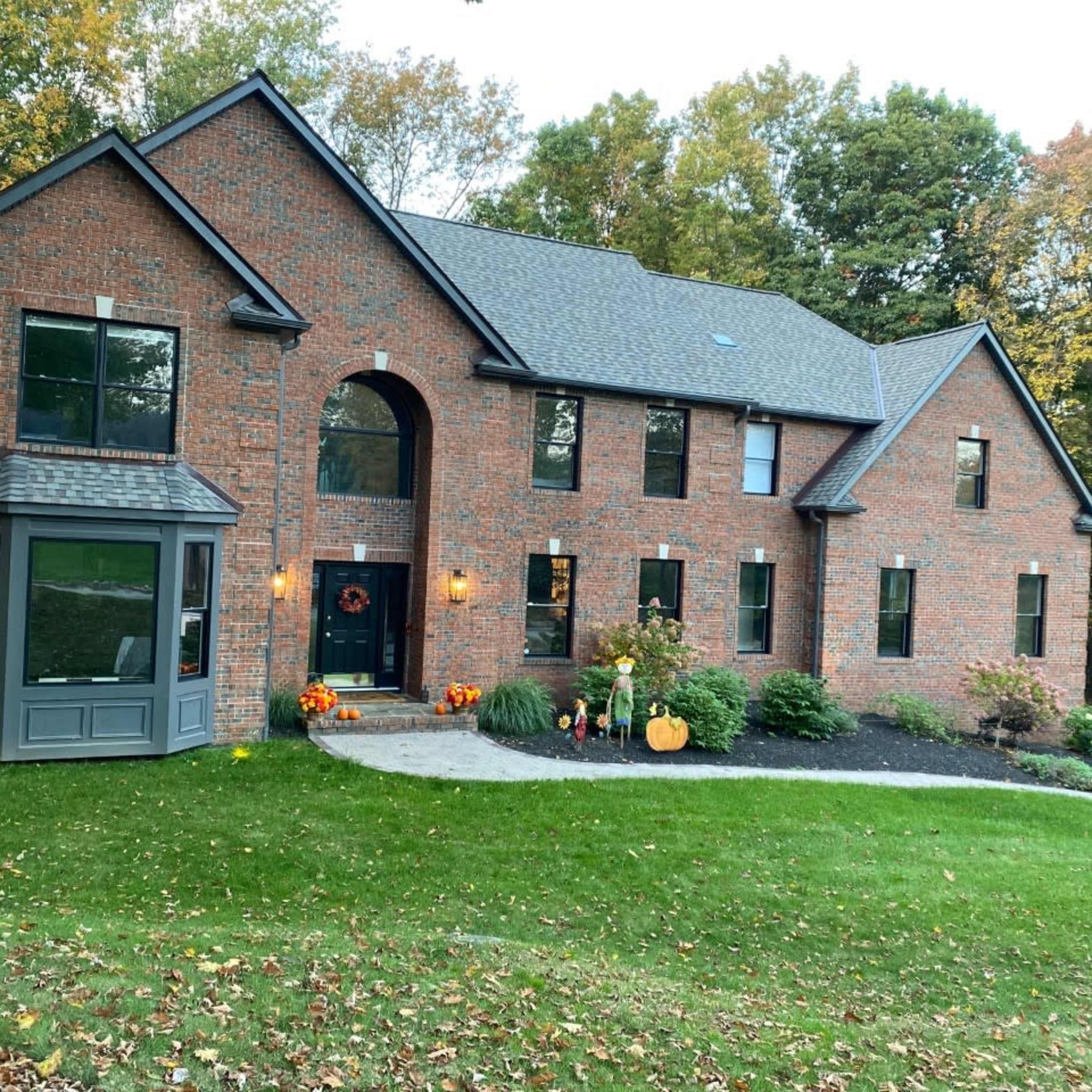A two-story brick house with large windows is surrounded by a well-kept lawn and autumn decorations, including pumpkins and orange flowers.