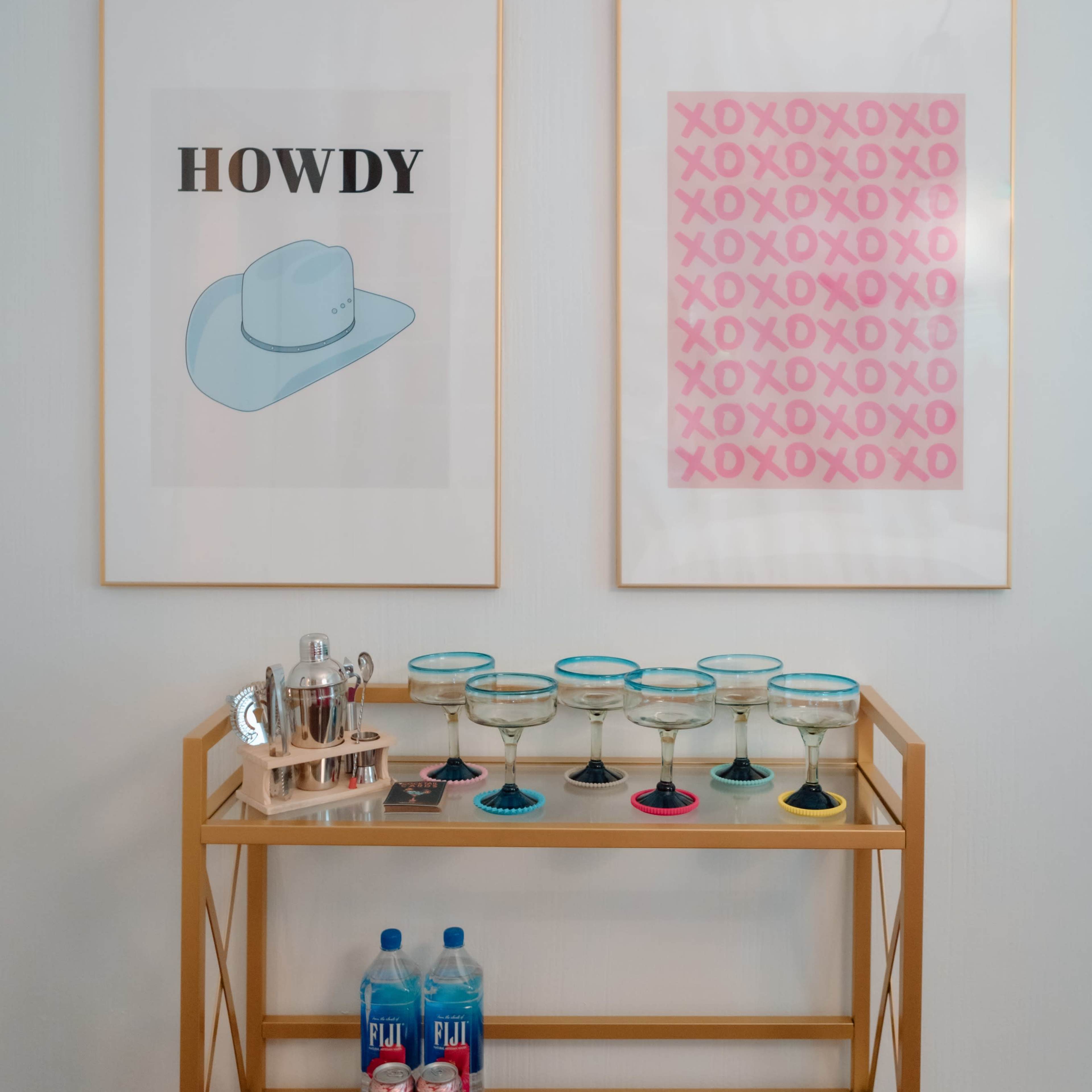 A gold bar cart displays six margarita glasses with colored rims, a cocktail shaker, and two bottles of flavored water, situated beneath two framed art pieces featuring colorful designs and text.