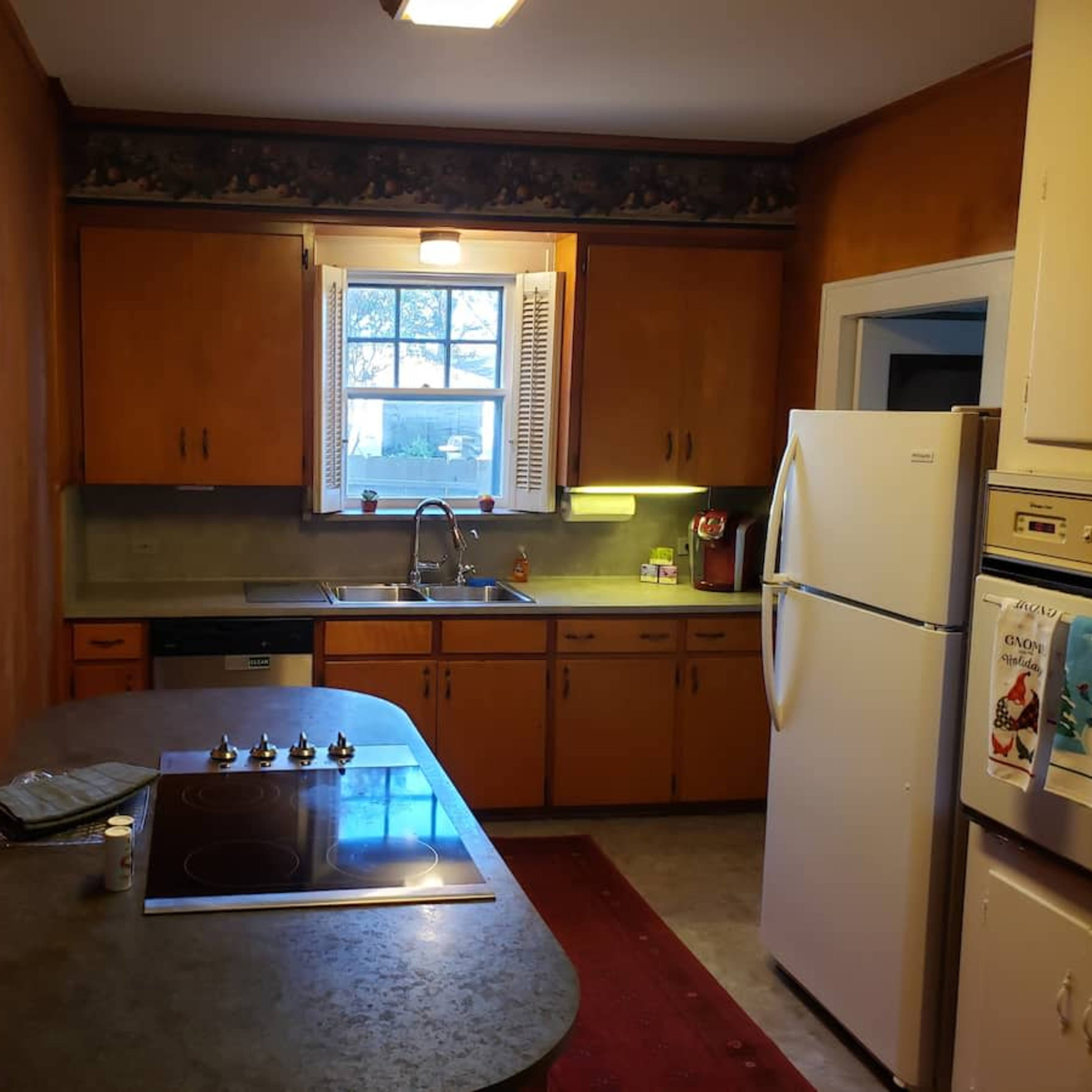 The kitchen features wooden cabinets, a sink beneath a window, a countertop with a cooktop, and a refrigerator against an adjacent wall.