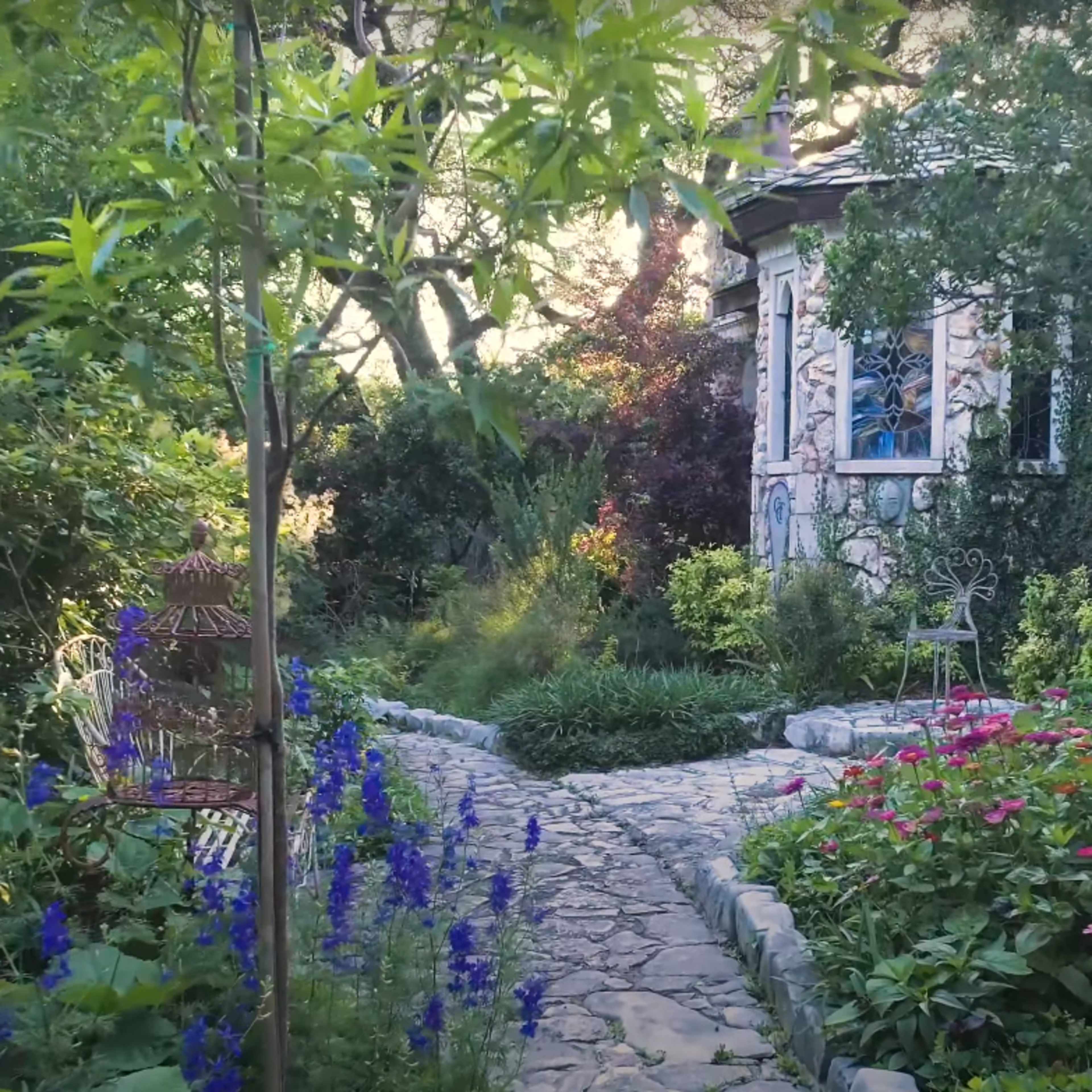 A stone path winds through a lush garden filled with colorful flowers and greenery, leading to a small house.