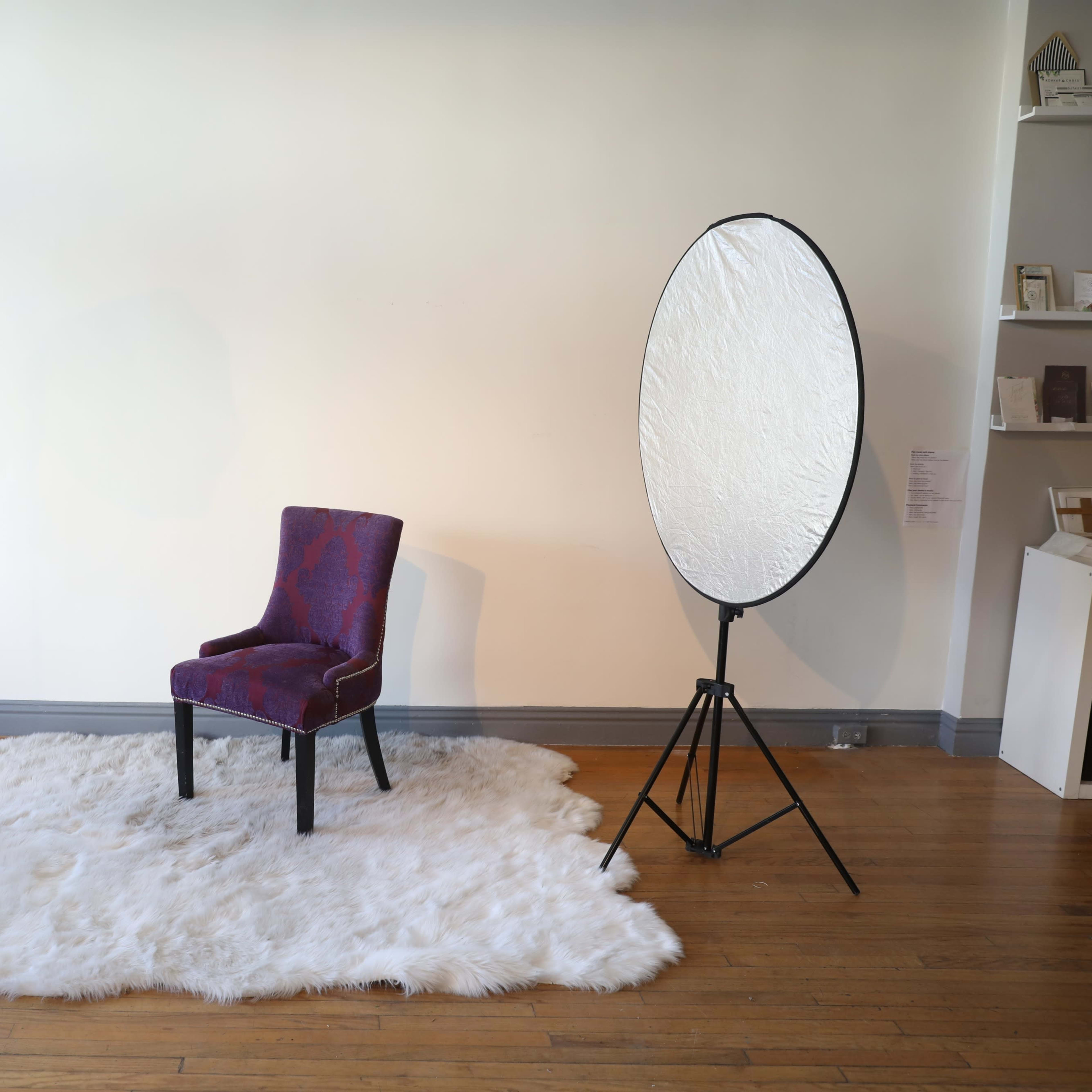 A studio setup featuring a patterned chair placed on a fluffy white rug, illuminated by a softbox light and a large reflector.