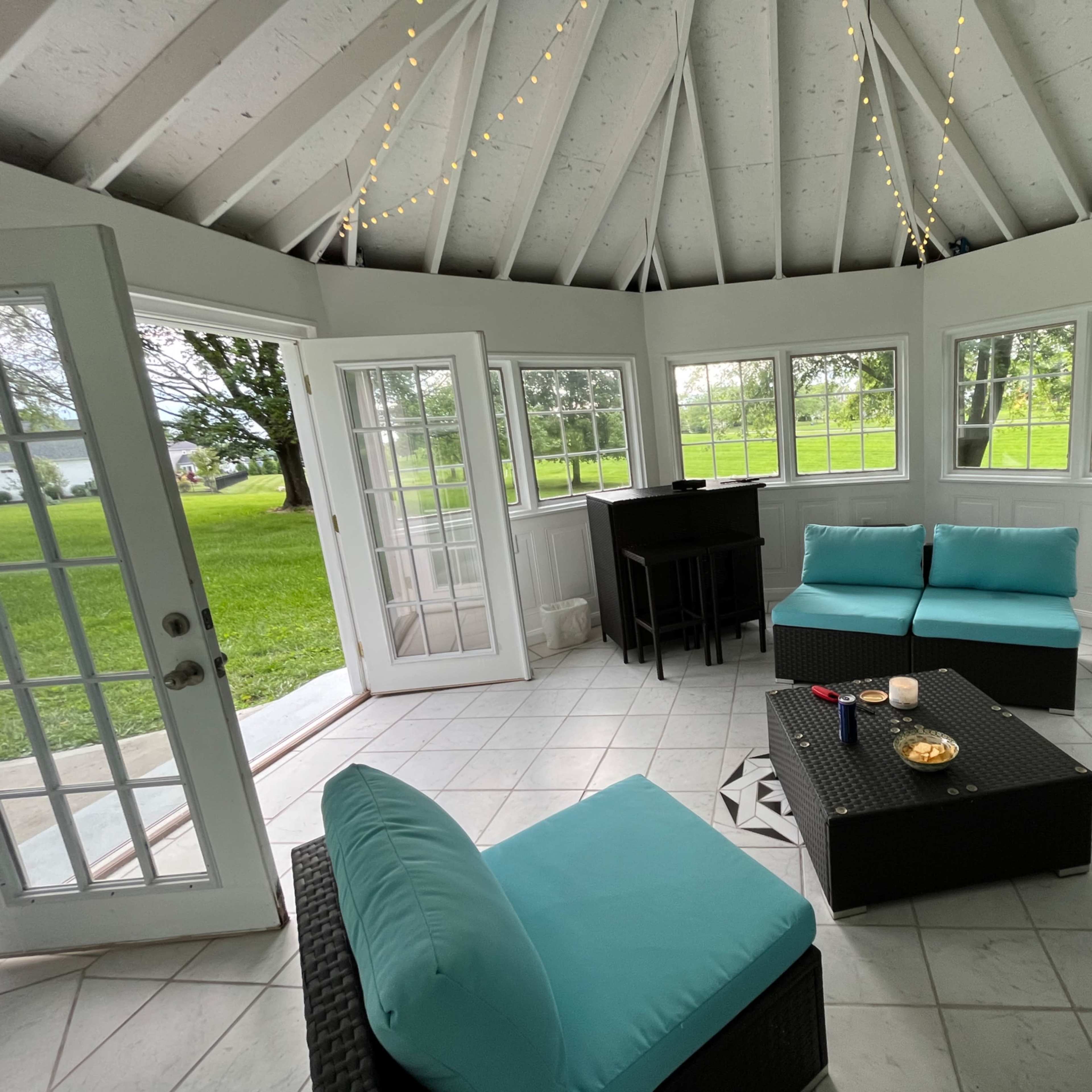 A gazebo with a tiled floor, featuring a seating area with turquoise cushions and a small table, surrounded by large windows and doors.