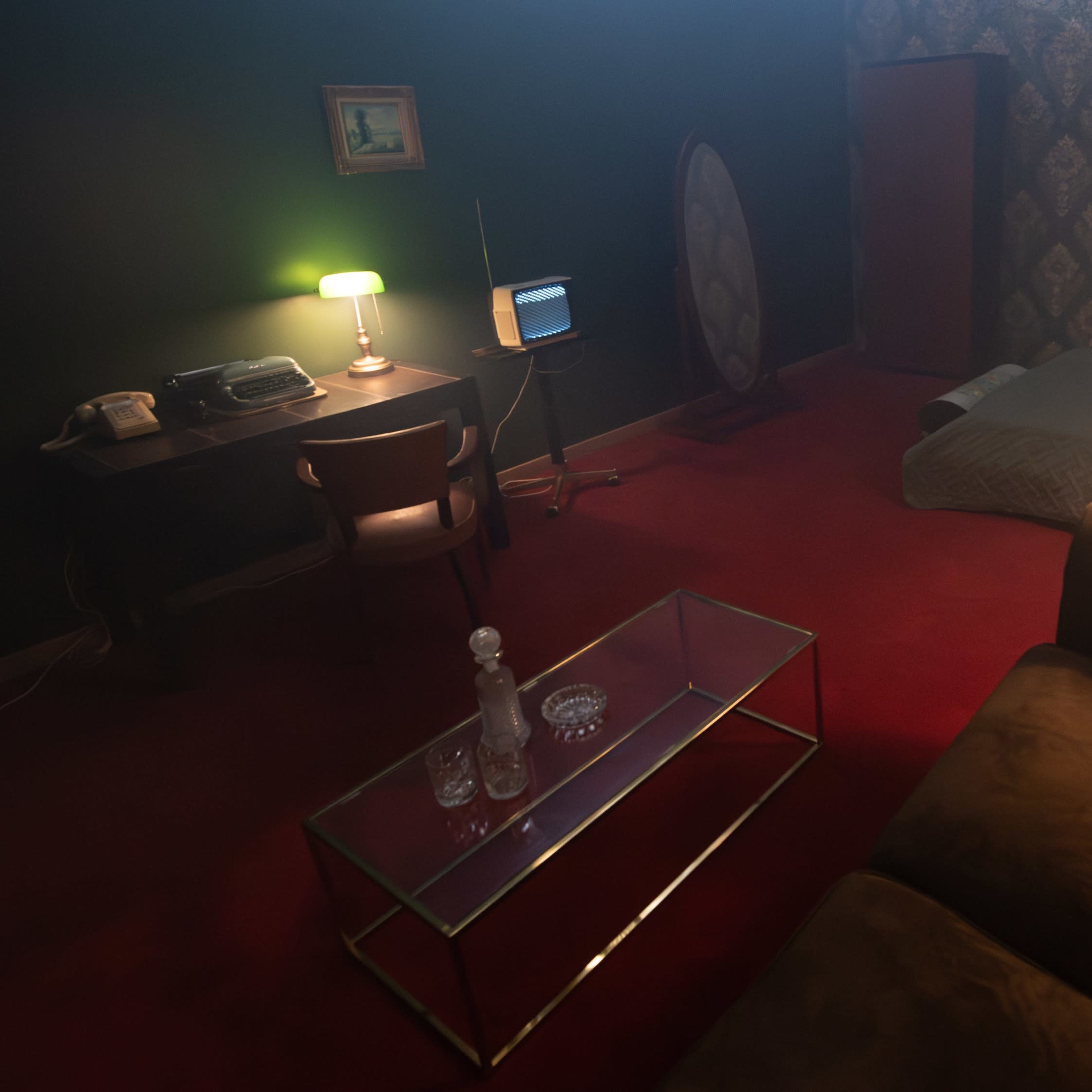 A dimly lit room with a bed, a vintage-style desk and chair, an old television, a lamp, and a glass coffee table with decanters.