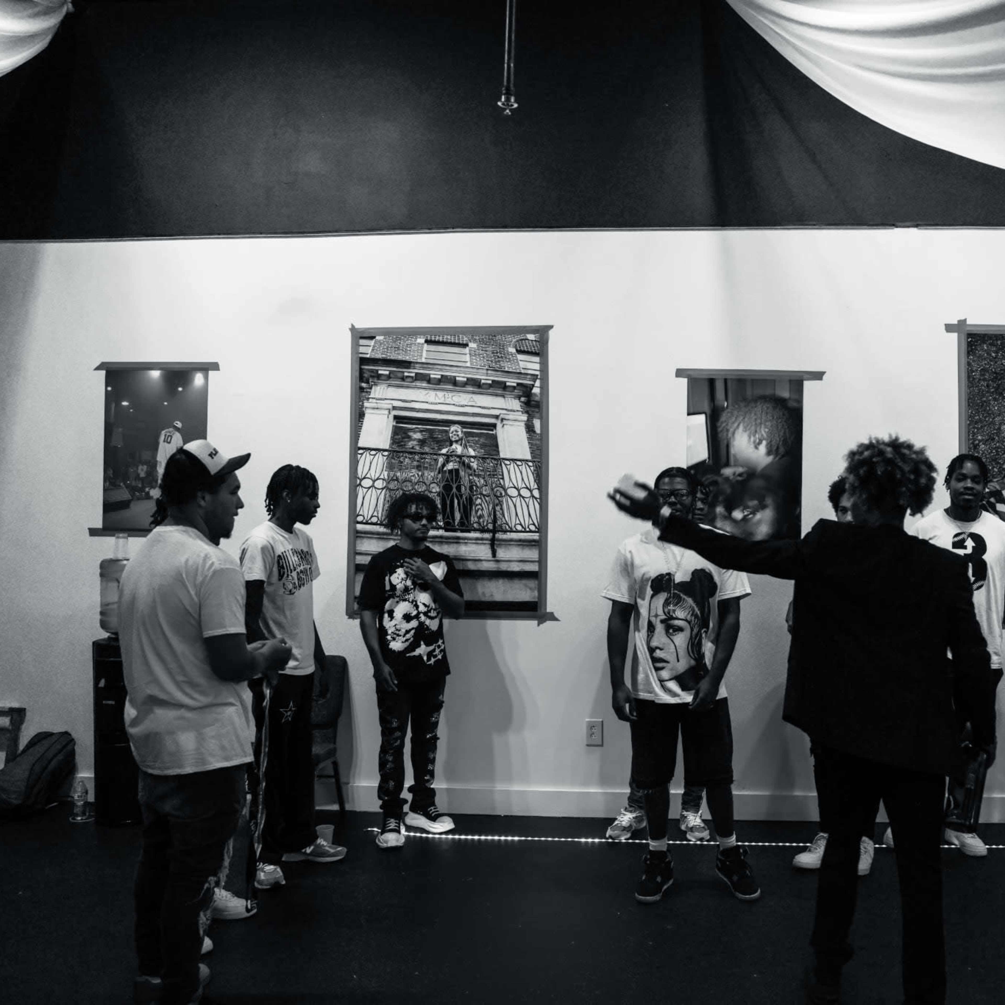 A group of people stands in a gallery space with framed artwork on the walls, while another person gestures toward them.