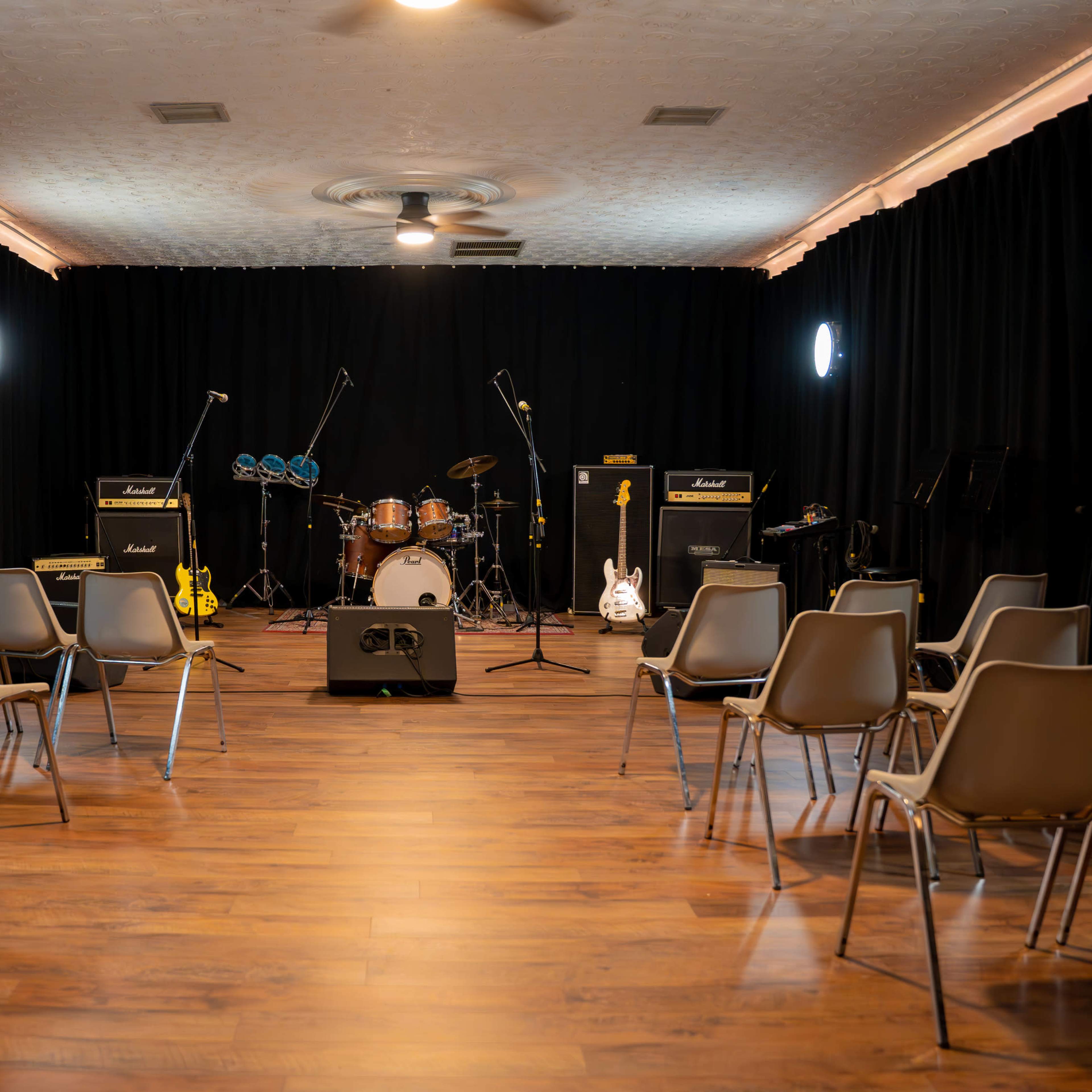 A small music performance space with rows of chairs facing a drum set and amplifiers against a black curtain backdrop.