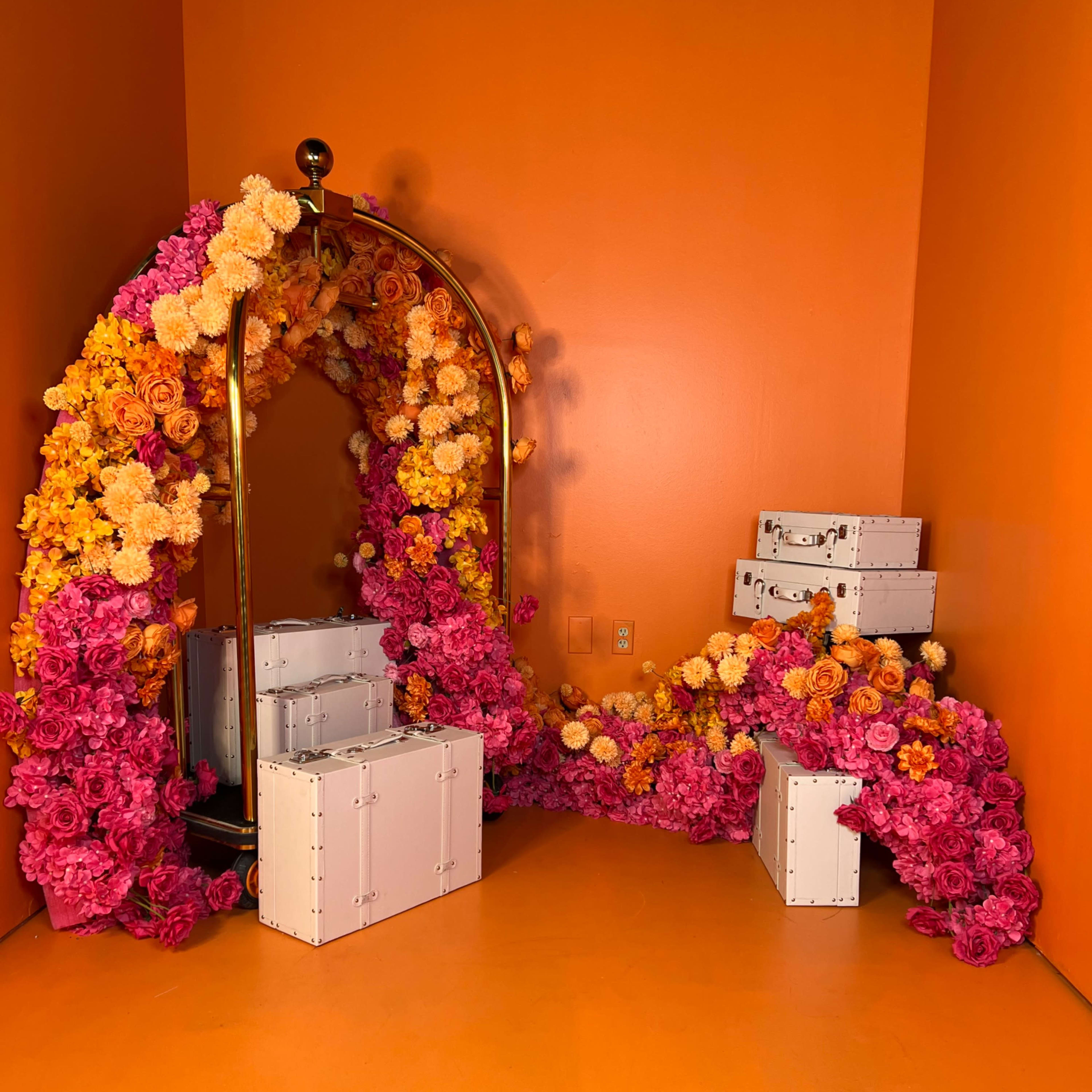 The scene features a vibrant orange wall adorned with an arch made of flowers in shades of pink and yellow, alongside stacked white suitcases.
