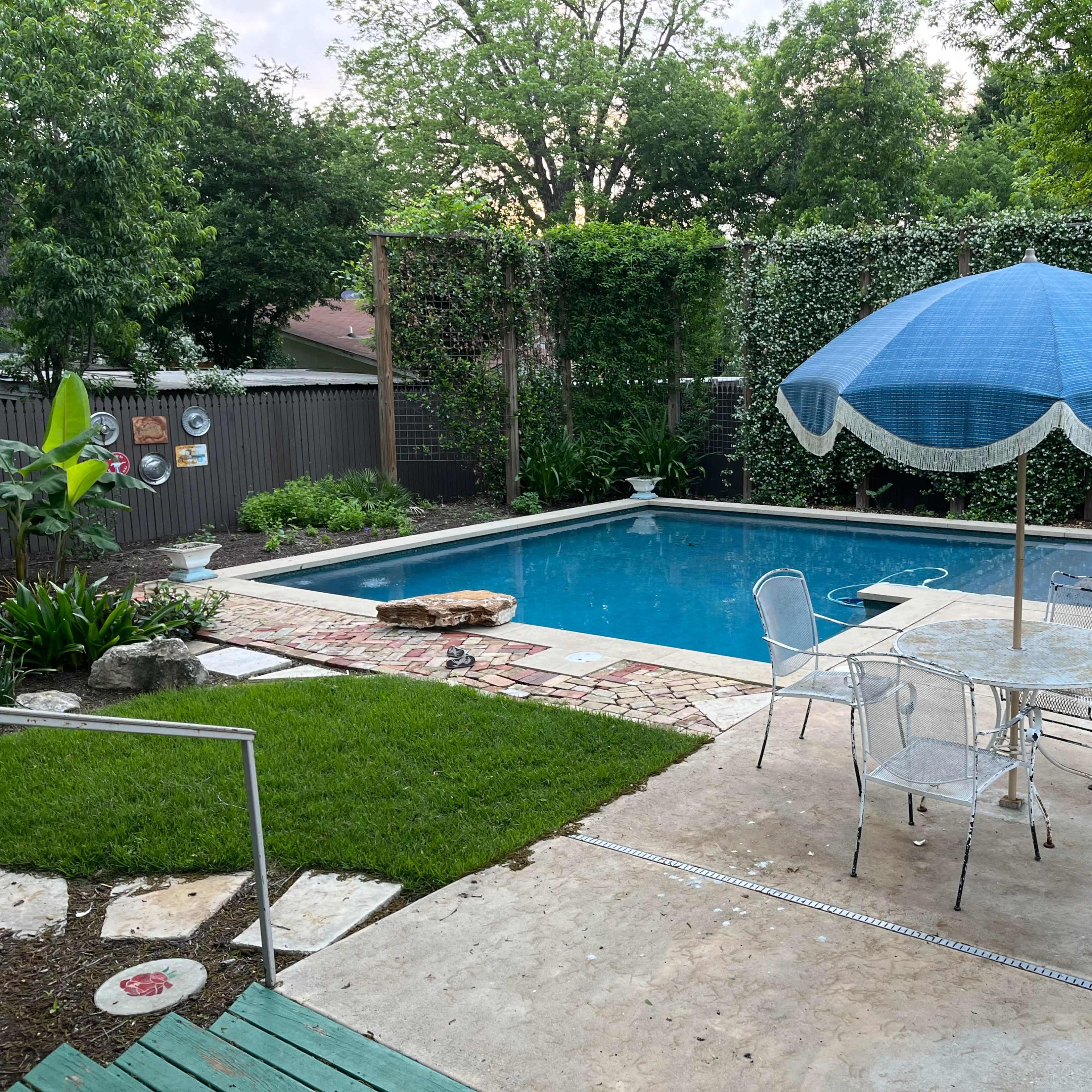 A backyard with a swimming pool, surrounded by greenery, a patio table with chairs, and a blue umbrella.
