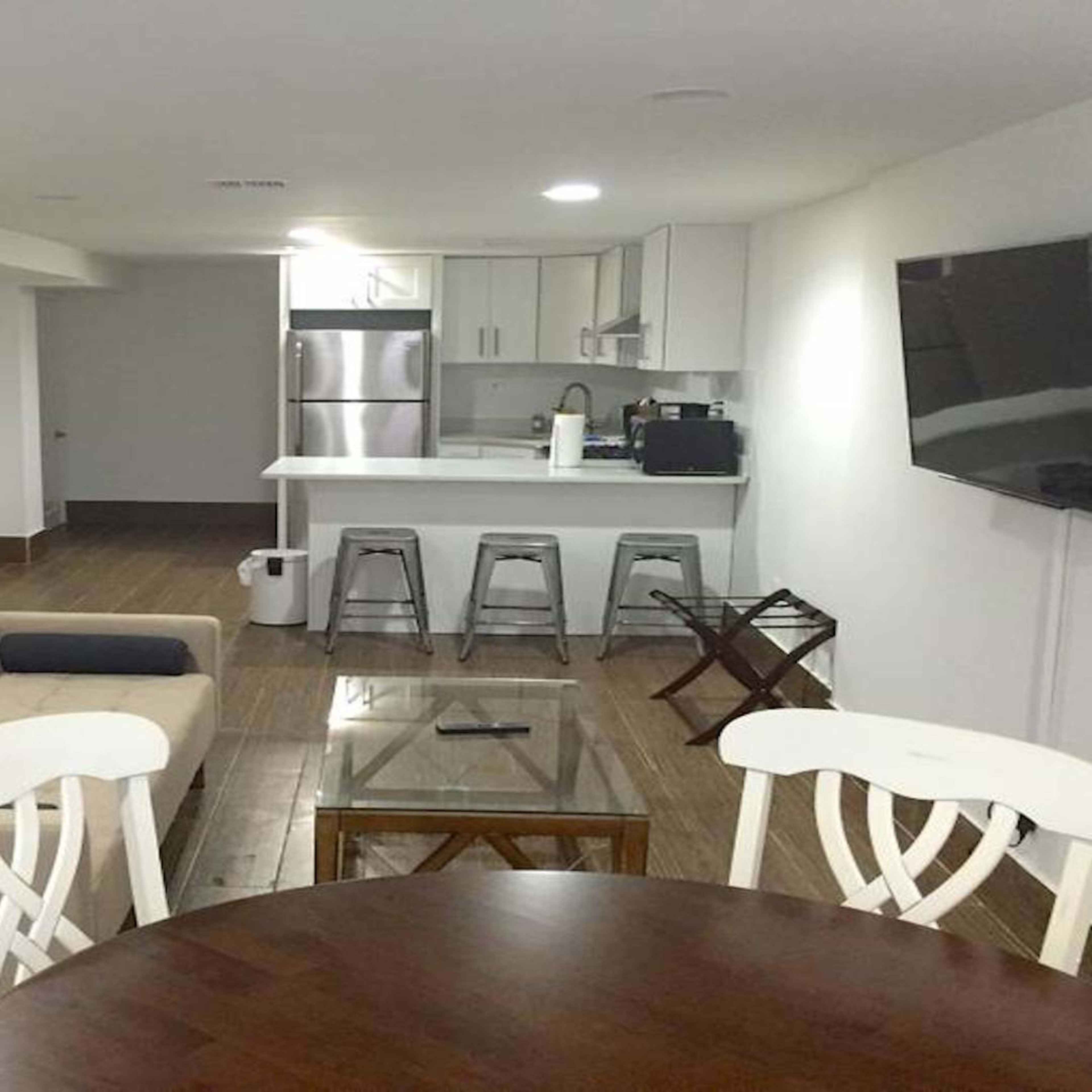 A modern, open-concept living space featuring a kitchen with stainless steel appliances, a beige sofa, a glass coffee table, a dining table with white chairs, and a mounted television.
