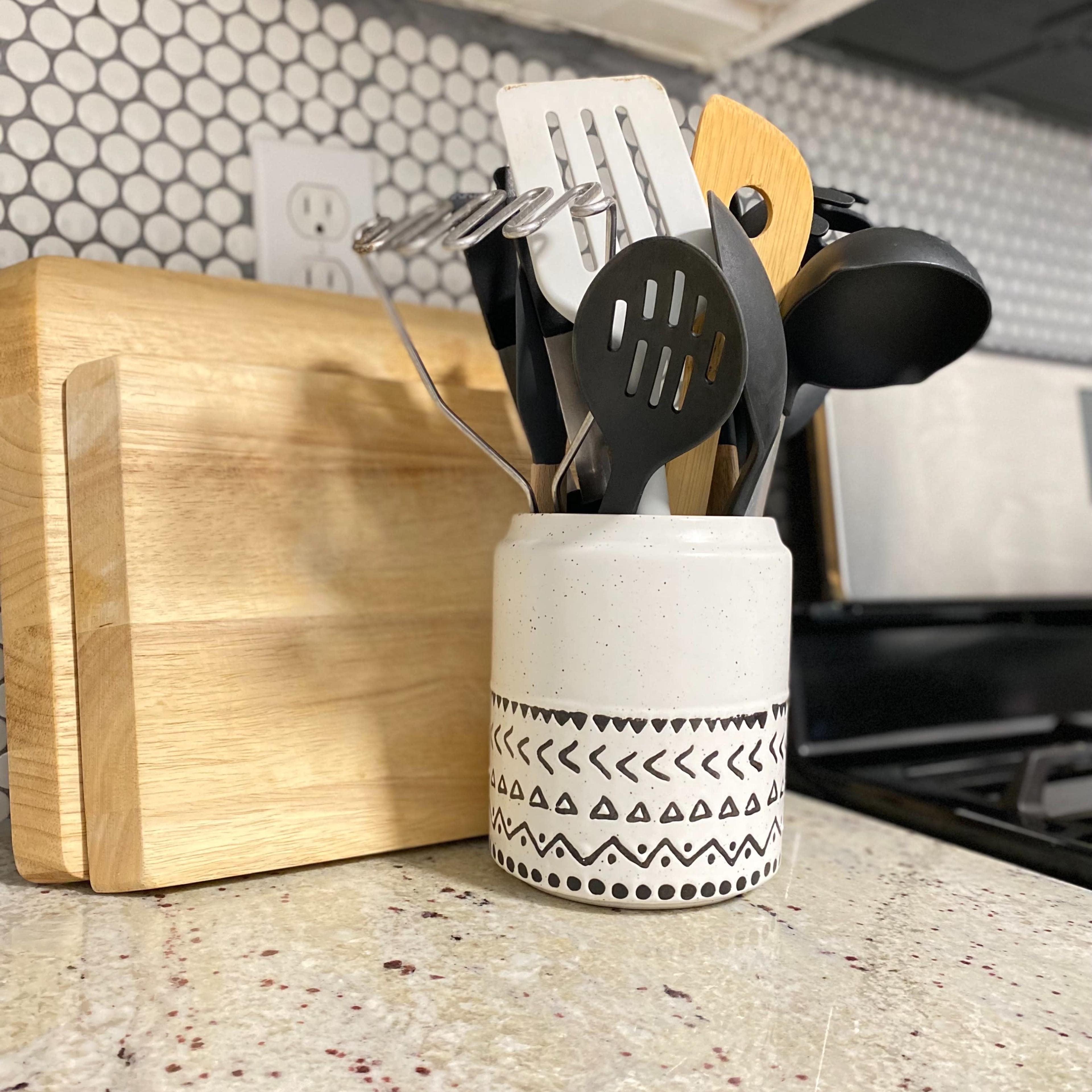 A white container with a patterned band holds various kitchen utensils next to a wooden cutting board on a speckled countertop.