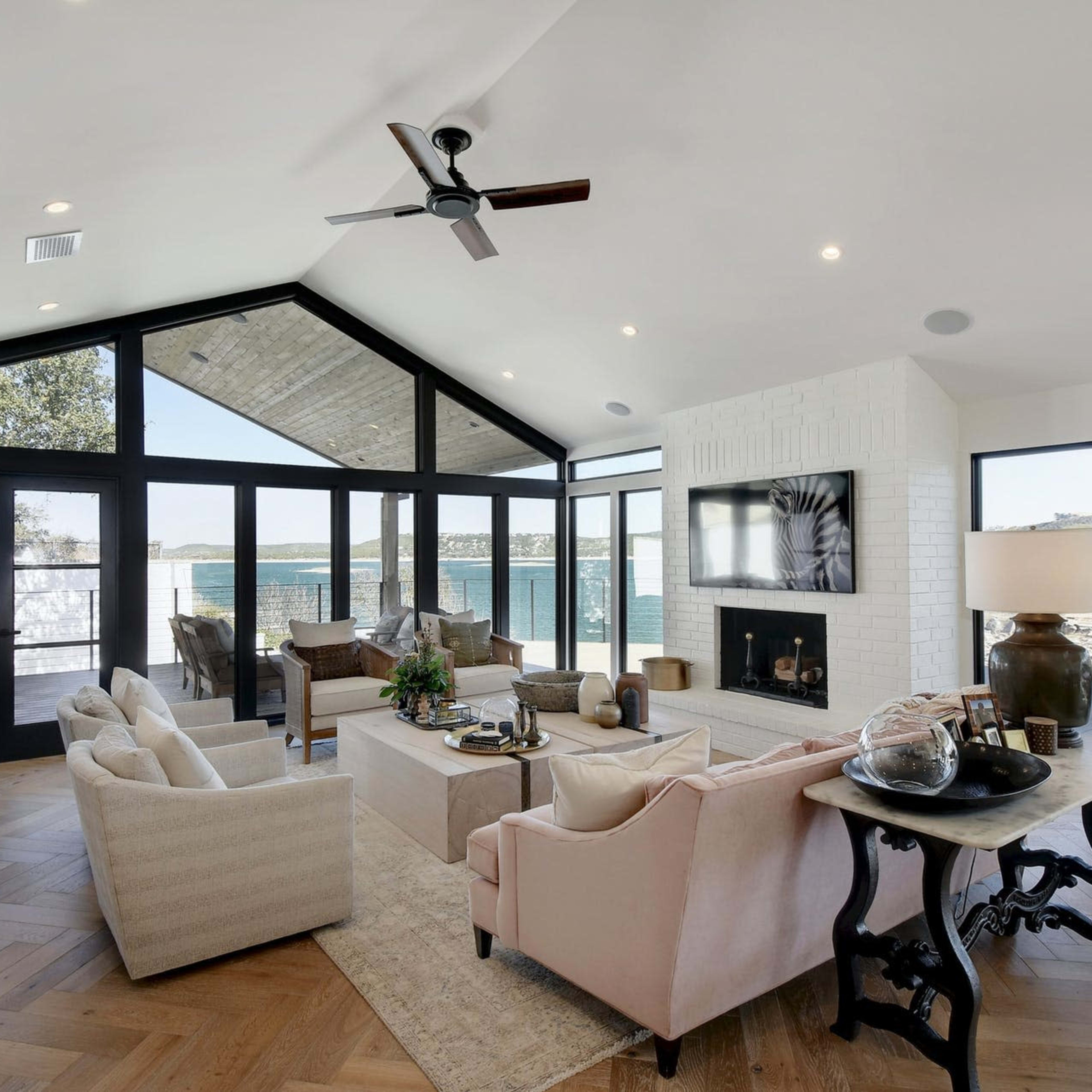 A spacious living room features large windows overlooking a body of water, with a mix of seating arrangements and a modern fireplace.