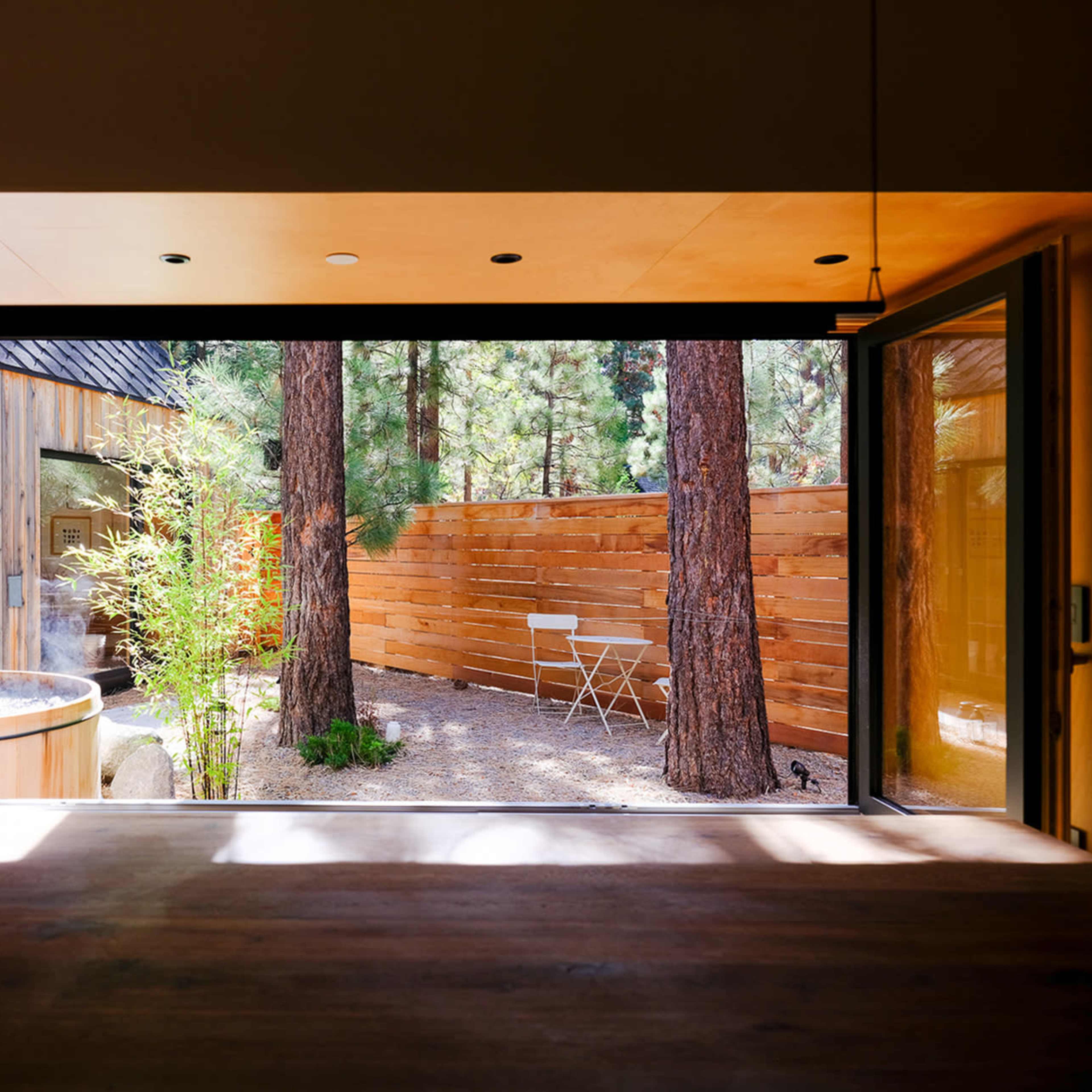 A modern interior space opening up to a serene outdoor area with a small table and hot tub surrounded by trees and wooden fencing.