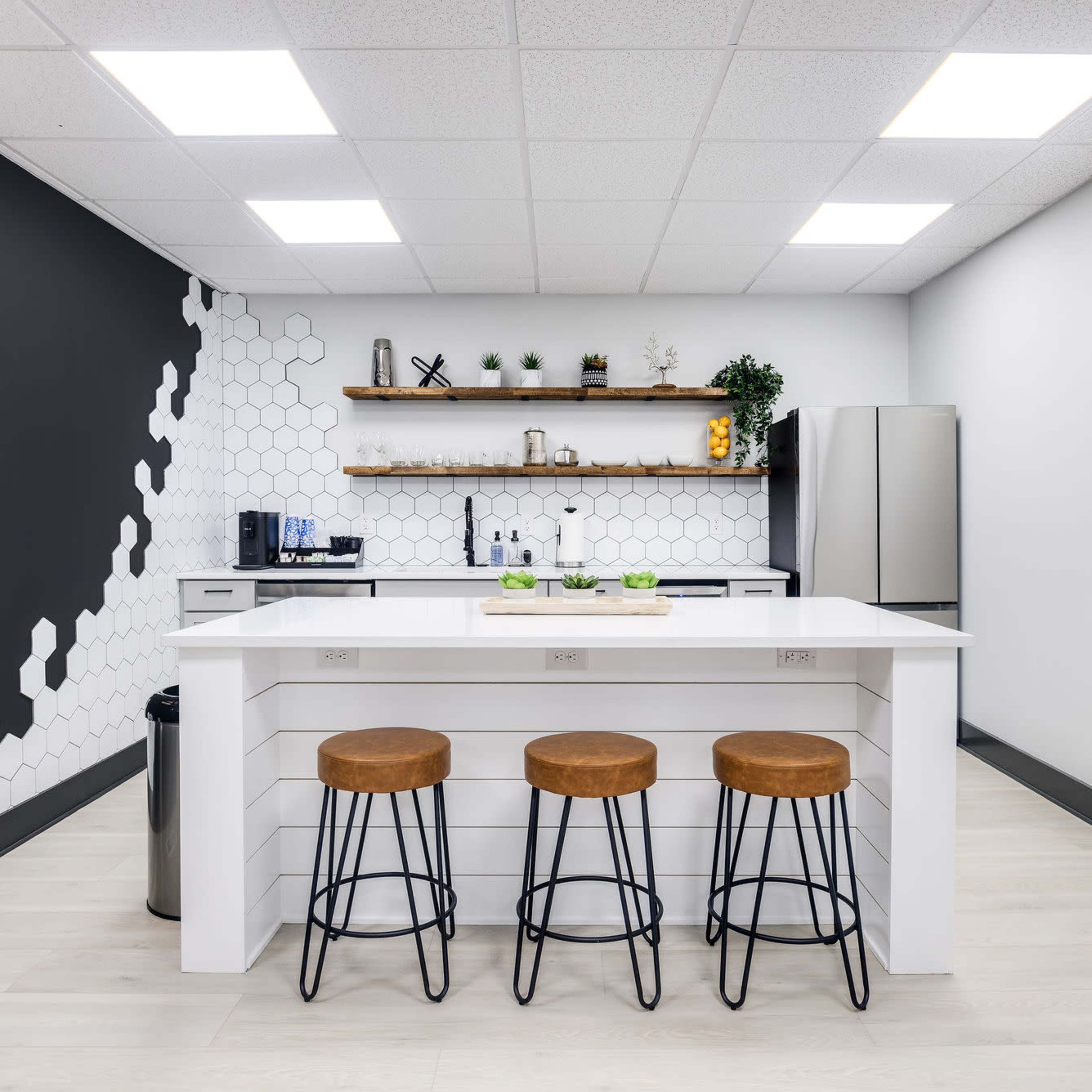 A modern kitchen with a white island, three metal stools, a hexagonal patterned wall, and a sleek silver refrigerator.