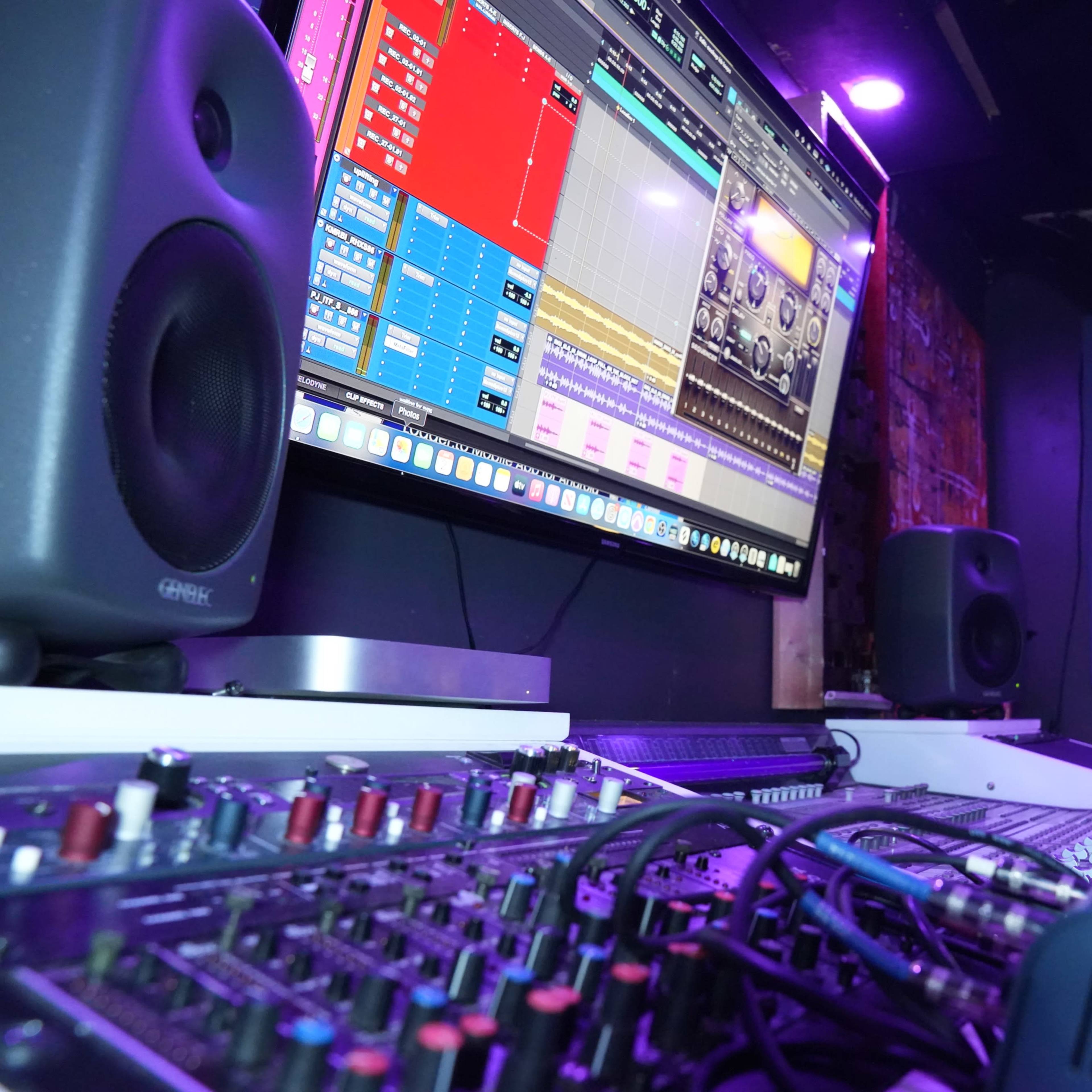 A music production workspace with a large monitor displaying software, audio equipment, and a colorful decorative figurine of a dog.
