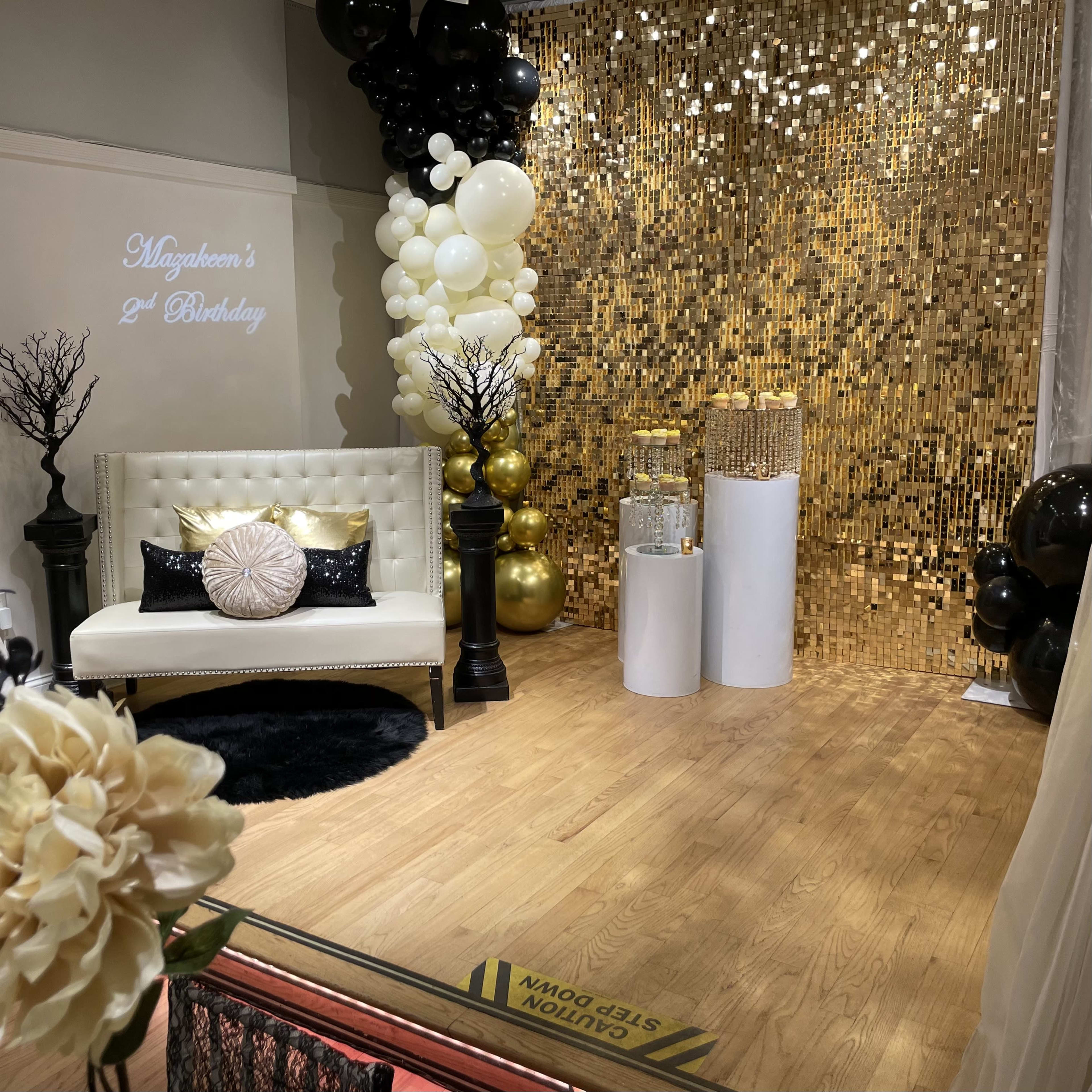 The scene features a decorated event space for a second birthday celebration, with a gold backdrop, a white sofa, and black and gold balloons.