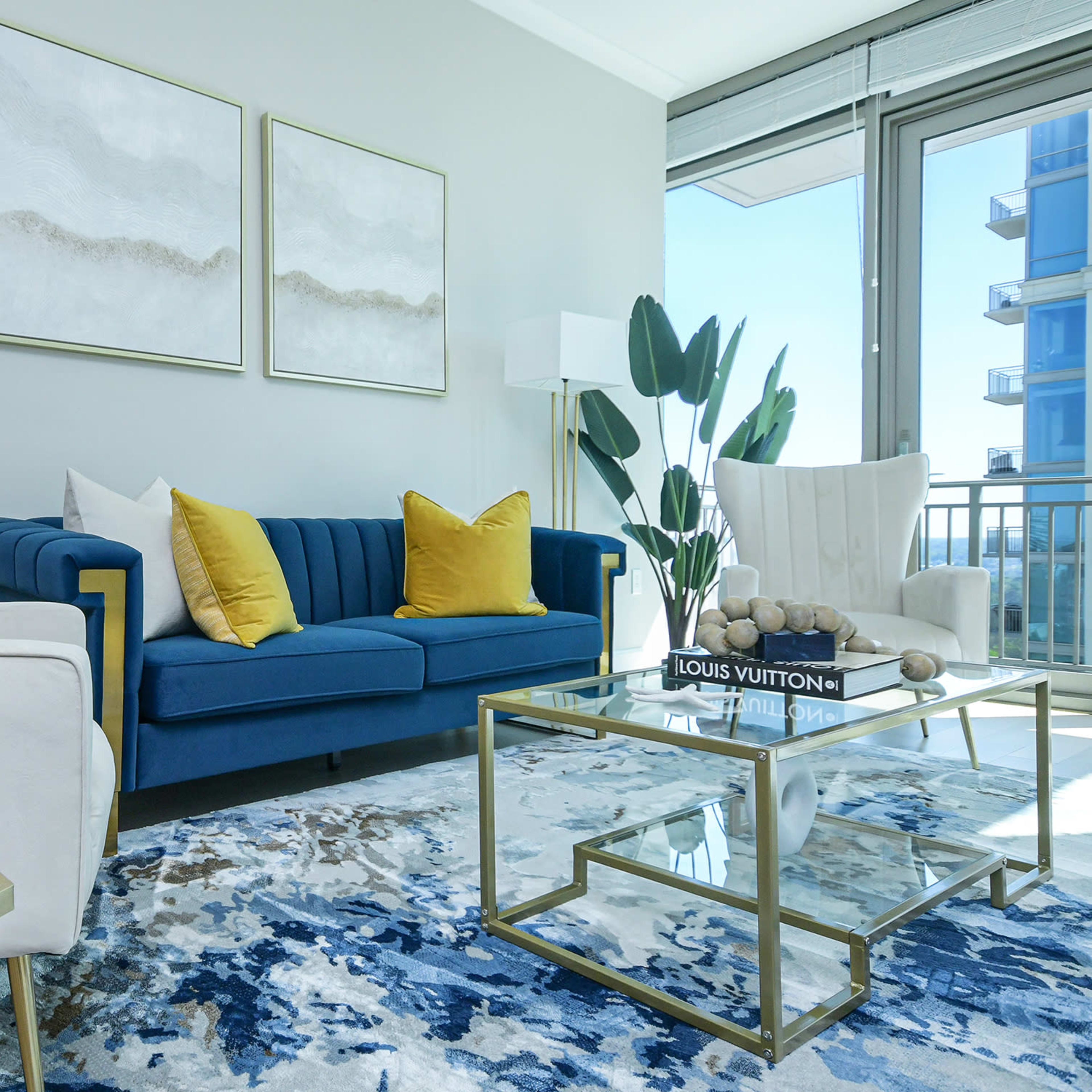 A modern living room with a blue sofa, two white armchairs, a glass coffee table, and decorative plants, all set against a light gray wall.