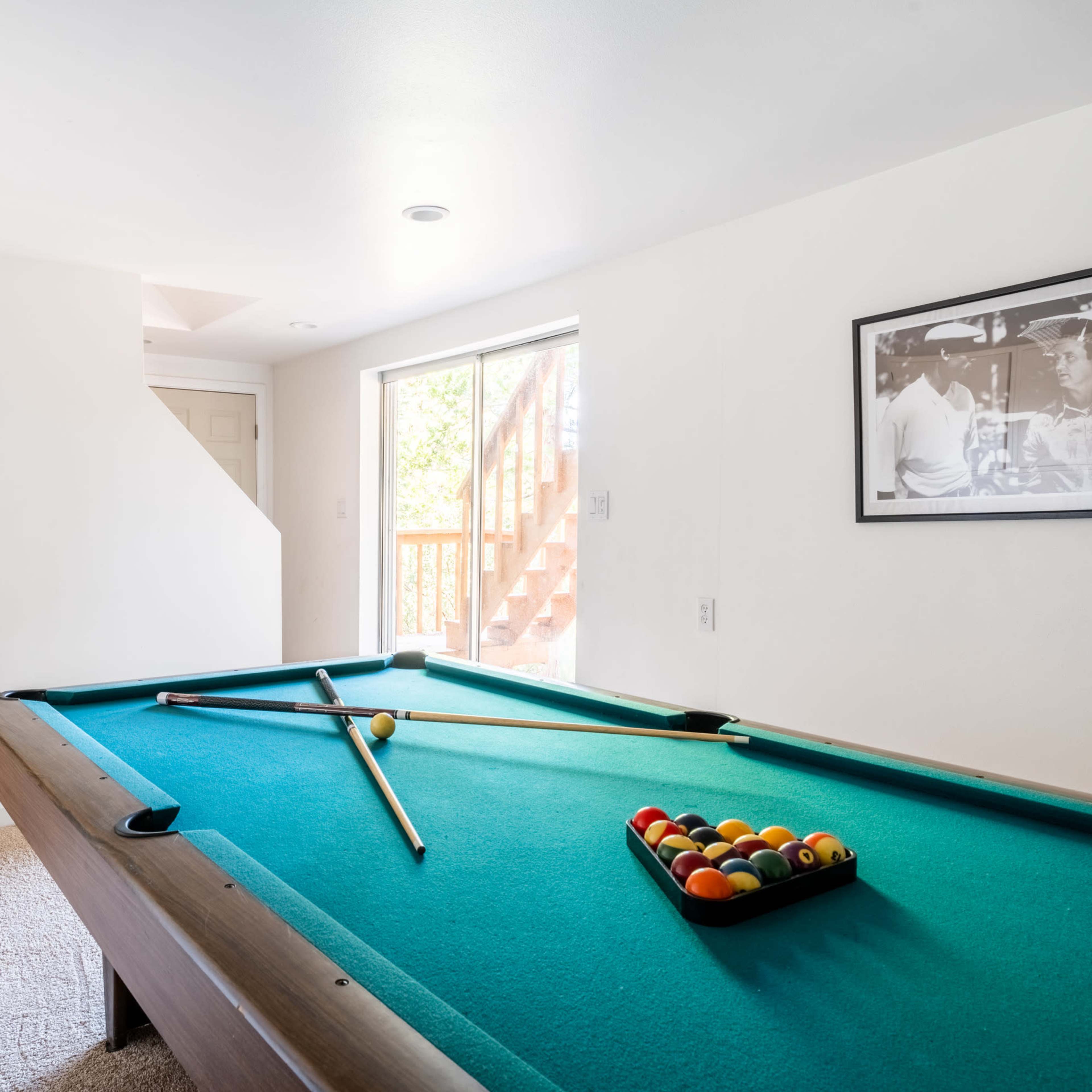 A billiard table with a triangle of colored balls is set in a bright room featuring two green bar stools and a photograph on the wall.