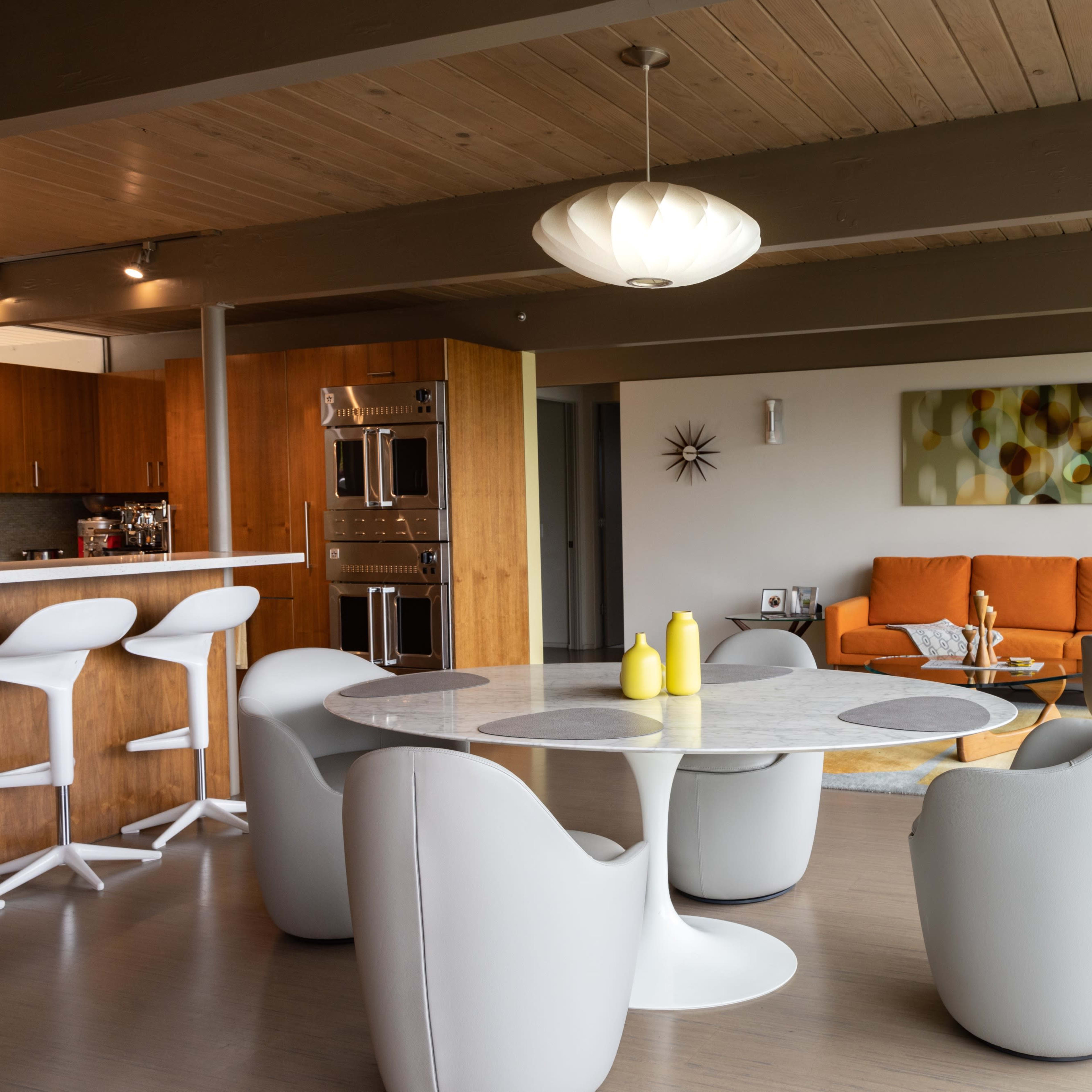 A modern living space features a round dining table with white chairs, an orange couch, and large windows allowing natural light to fill the room.