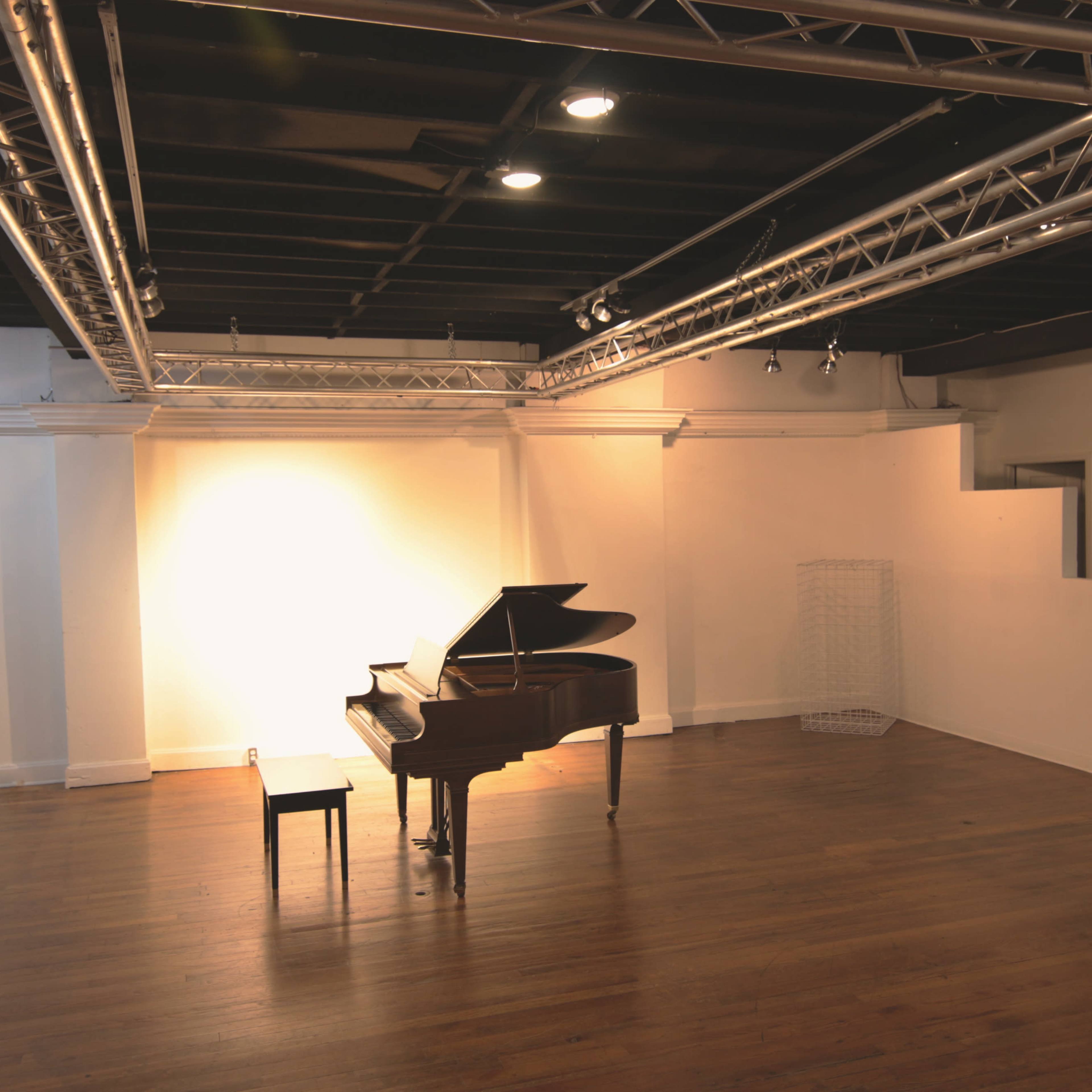 An empty room features a grand piano and a single chair under overhead lighting.