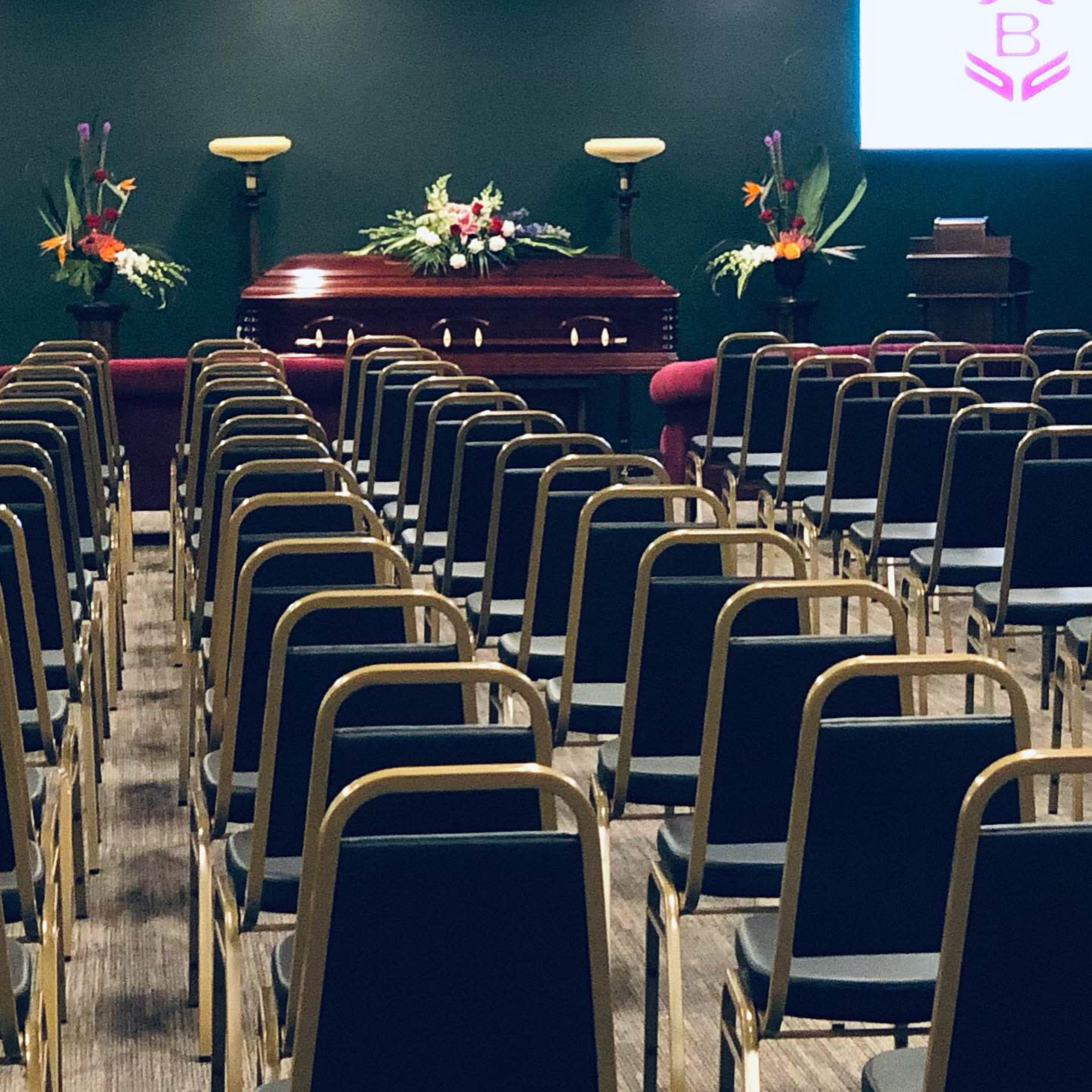 A funeral service setting with rows of chairs facing a casket adorned with flowers and two decorative floral arrangements on either side.