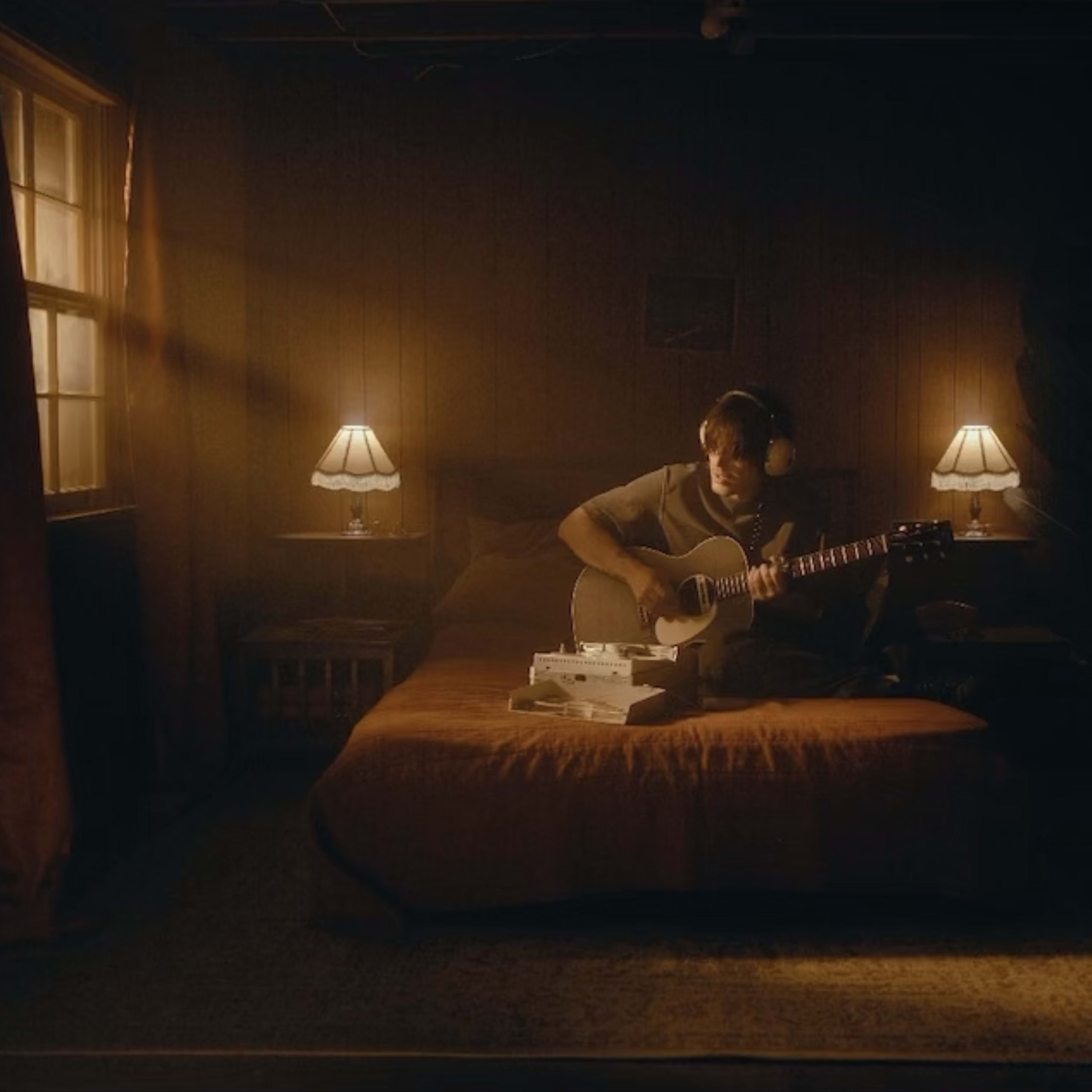 A musician sits on a bed in a dimly lit room, playing an acoustic guitar with a retro television and lamps on either side.