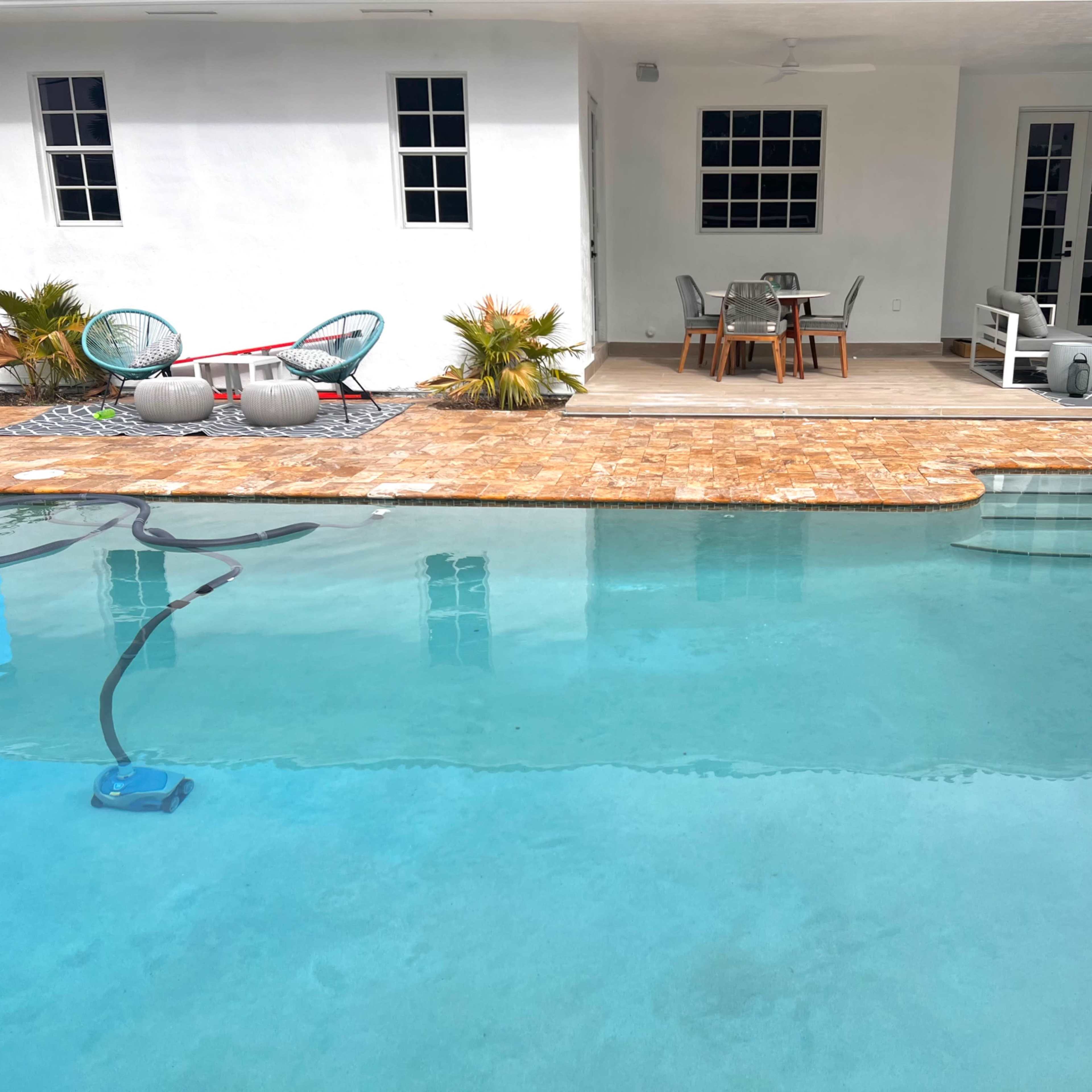 A clear pool is surrounded by a patio area with outdoor furniture, while a house with white walls and large windows is in the background.