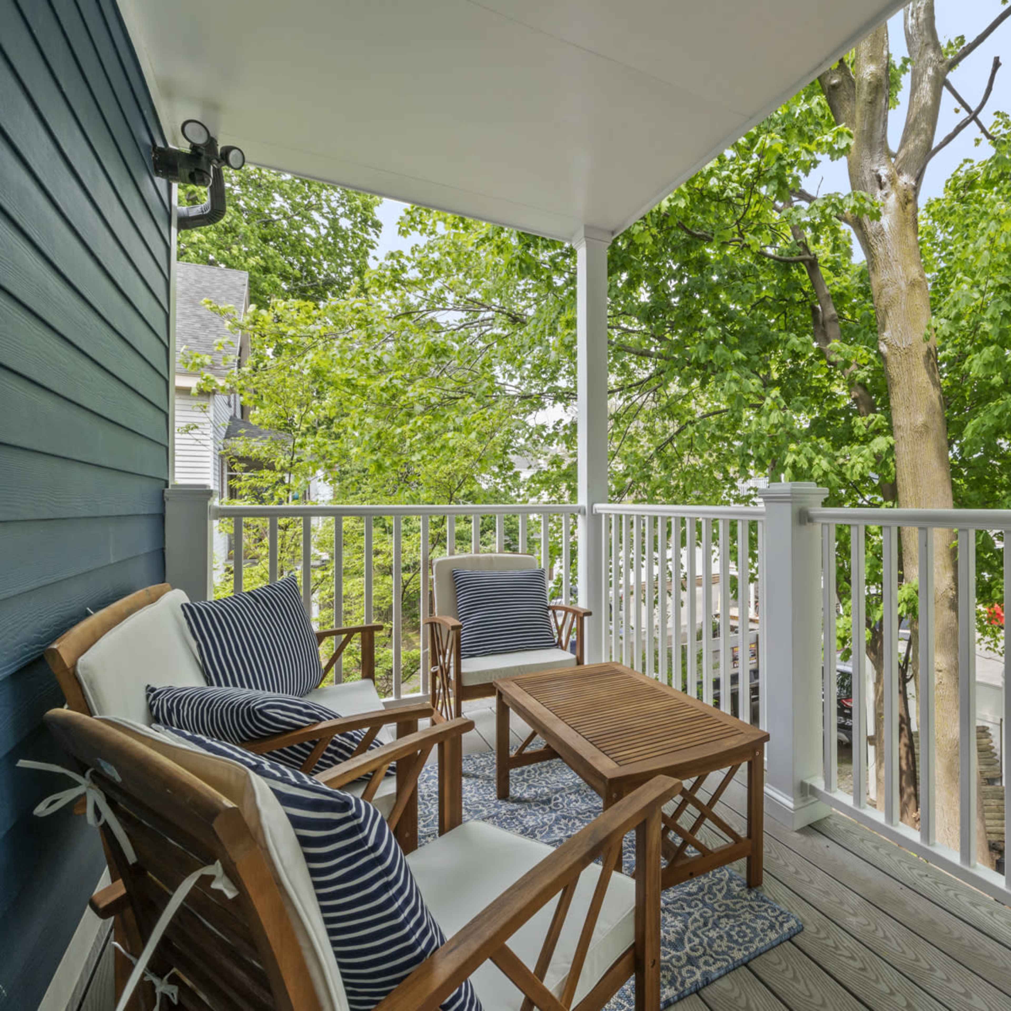 A porch features two wooden chairs and a small table, surrounded by greenery and a view of the street.