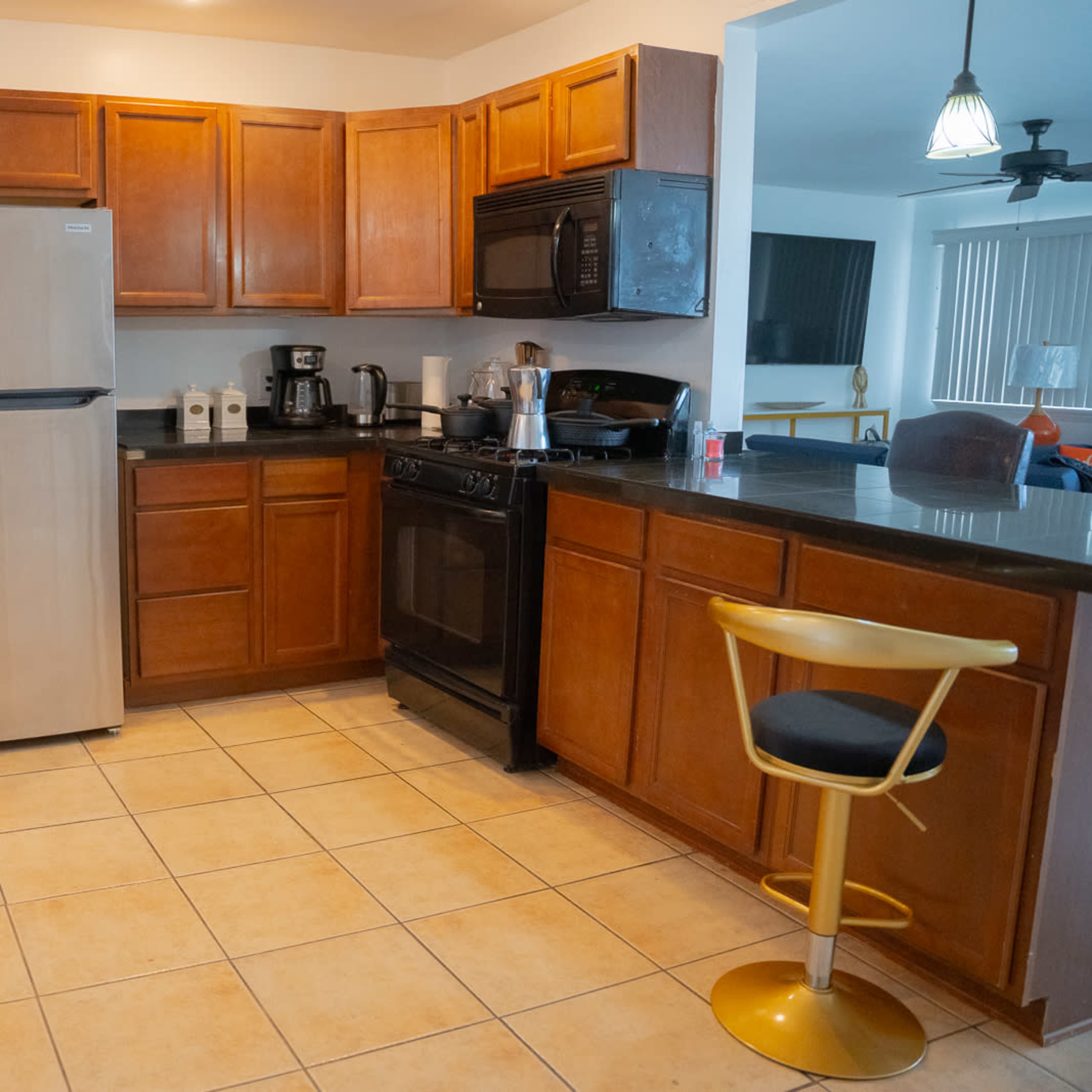 A kitchen with wooden cabinetry, stainless steel appliances, and a black countertop, featuring a stool at the end.
