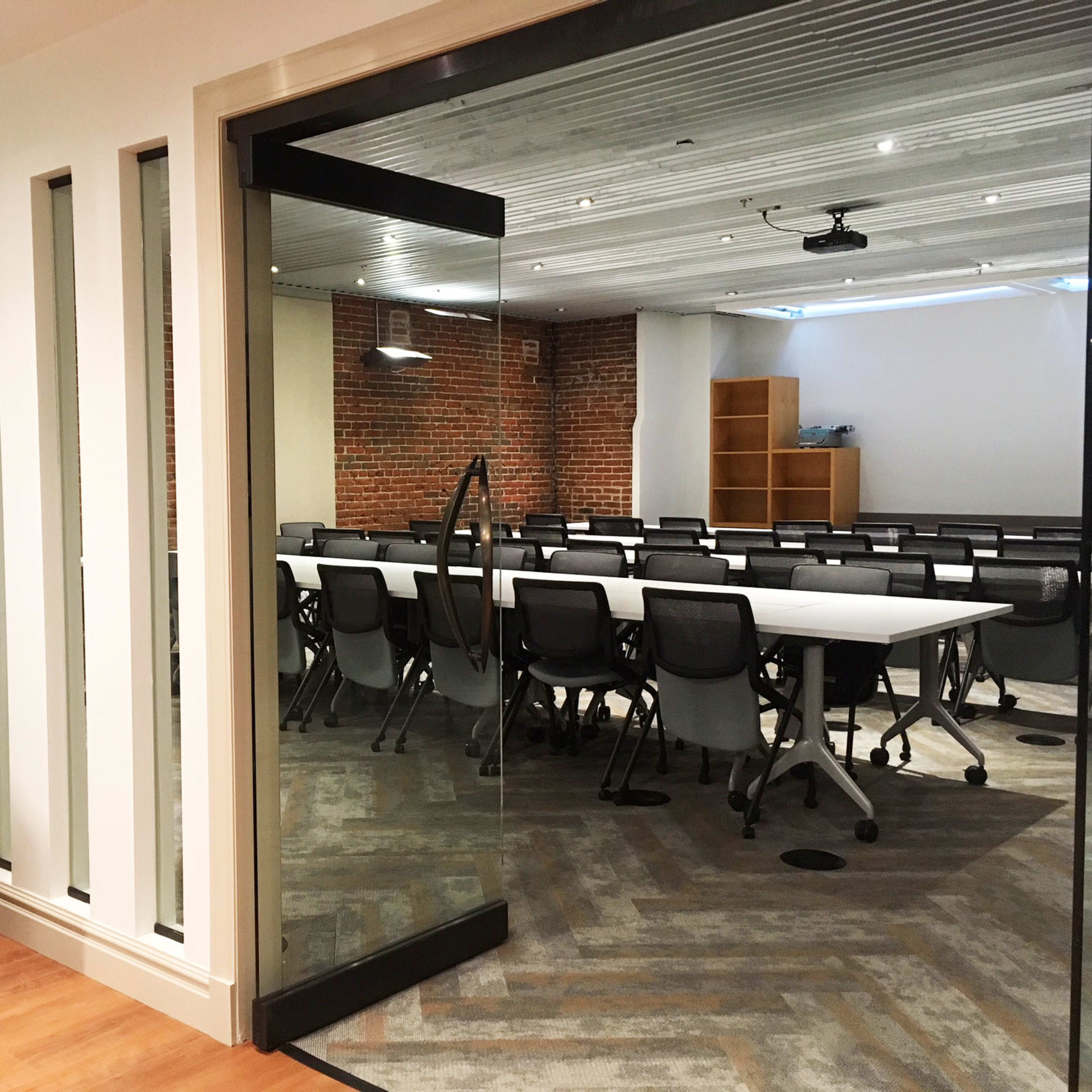A meeting room features a large, empty table surrounded by modern chairs, with a view through a glass door from a hallway.