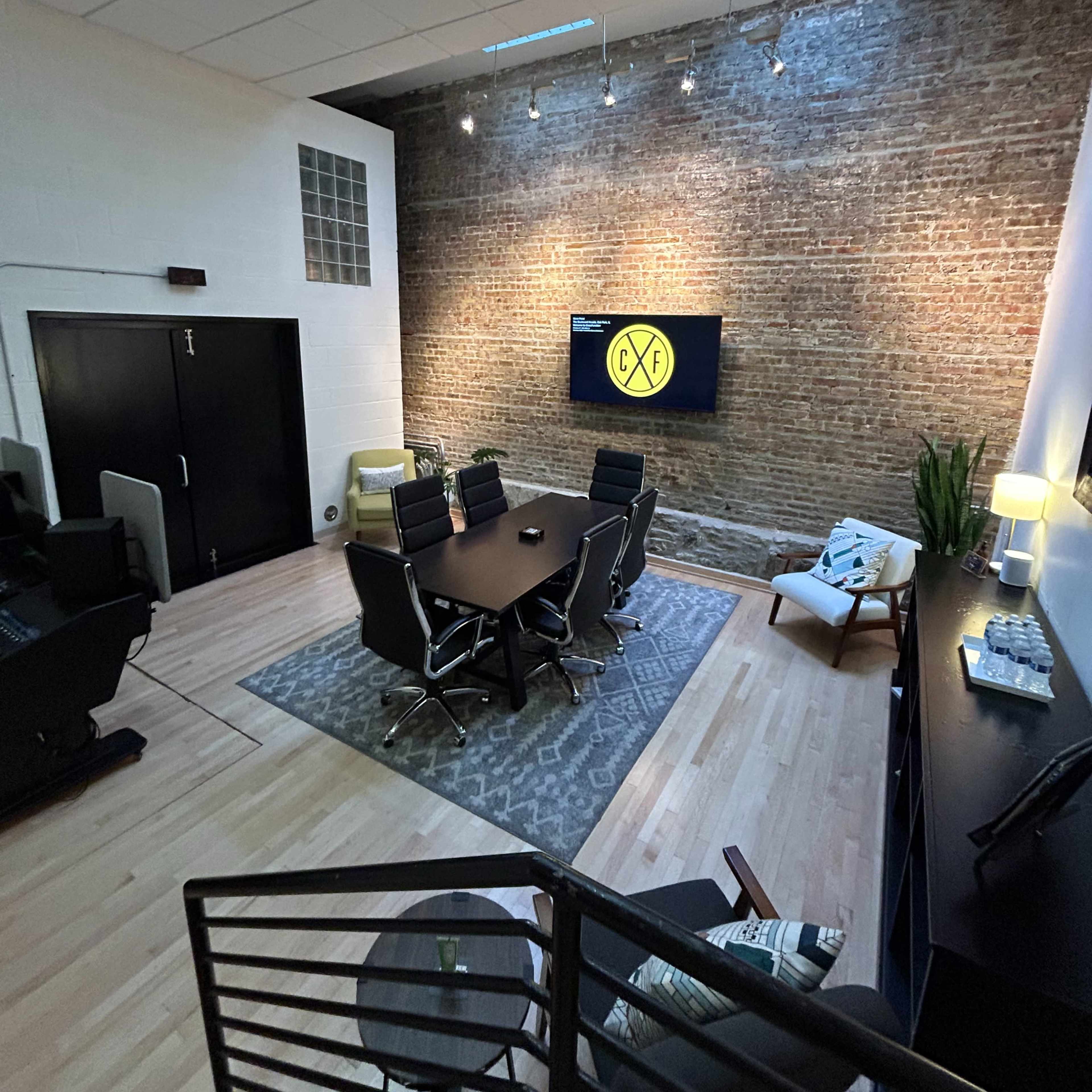 A modern office space featuring a conference table, ergonomic chairs, a large screen on a brick wall, and a seating area with a plant.