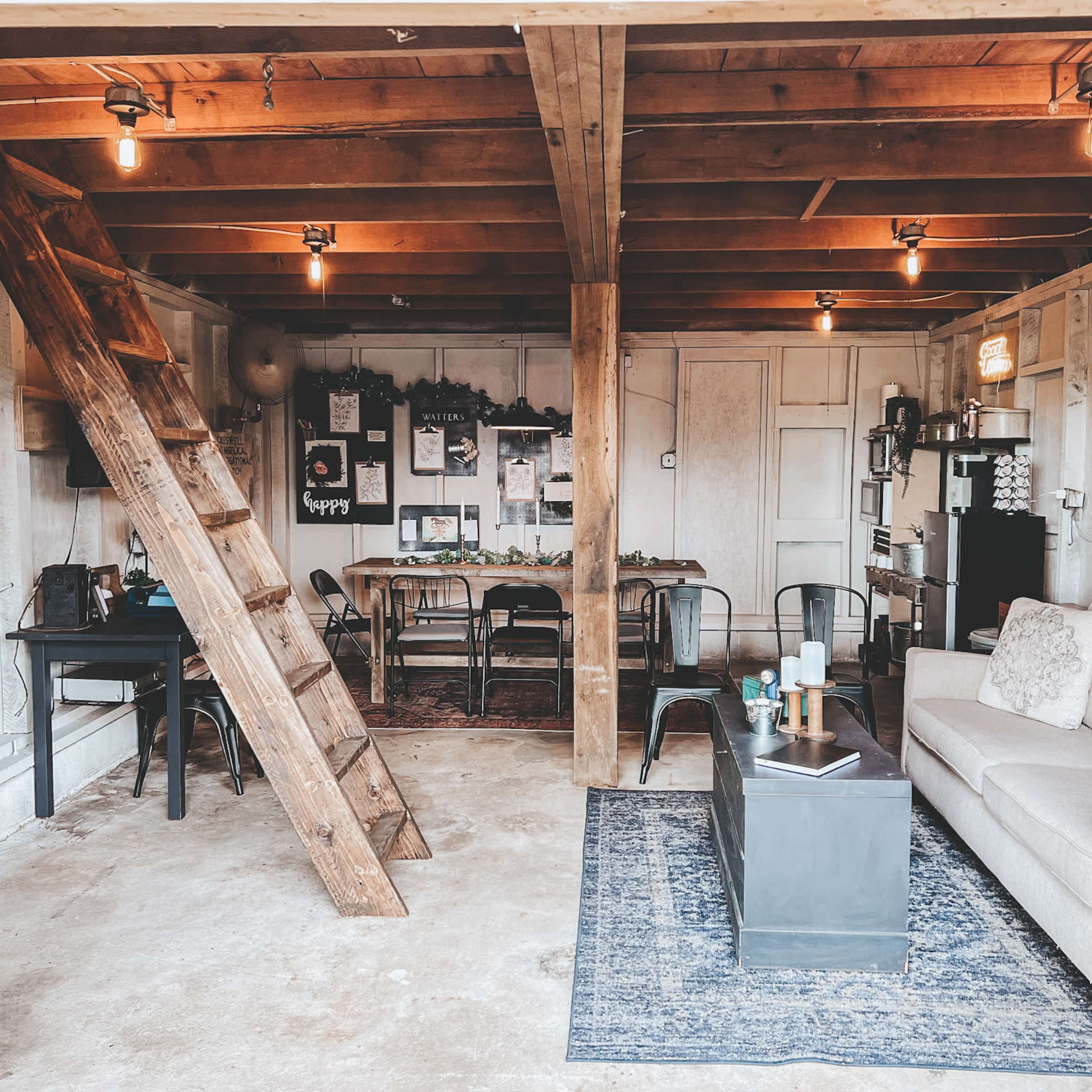 A garage interior converted into a cozy living space featuring a sofa, a table with chairs, a ladder leading to an upper area, and various decorative elements on the walls.