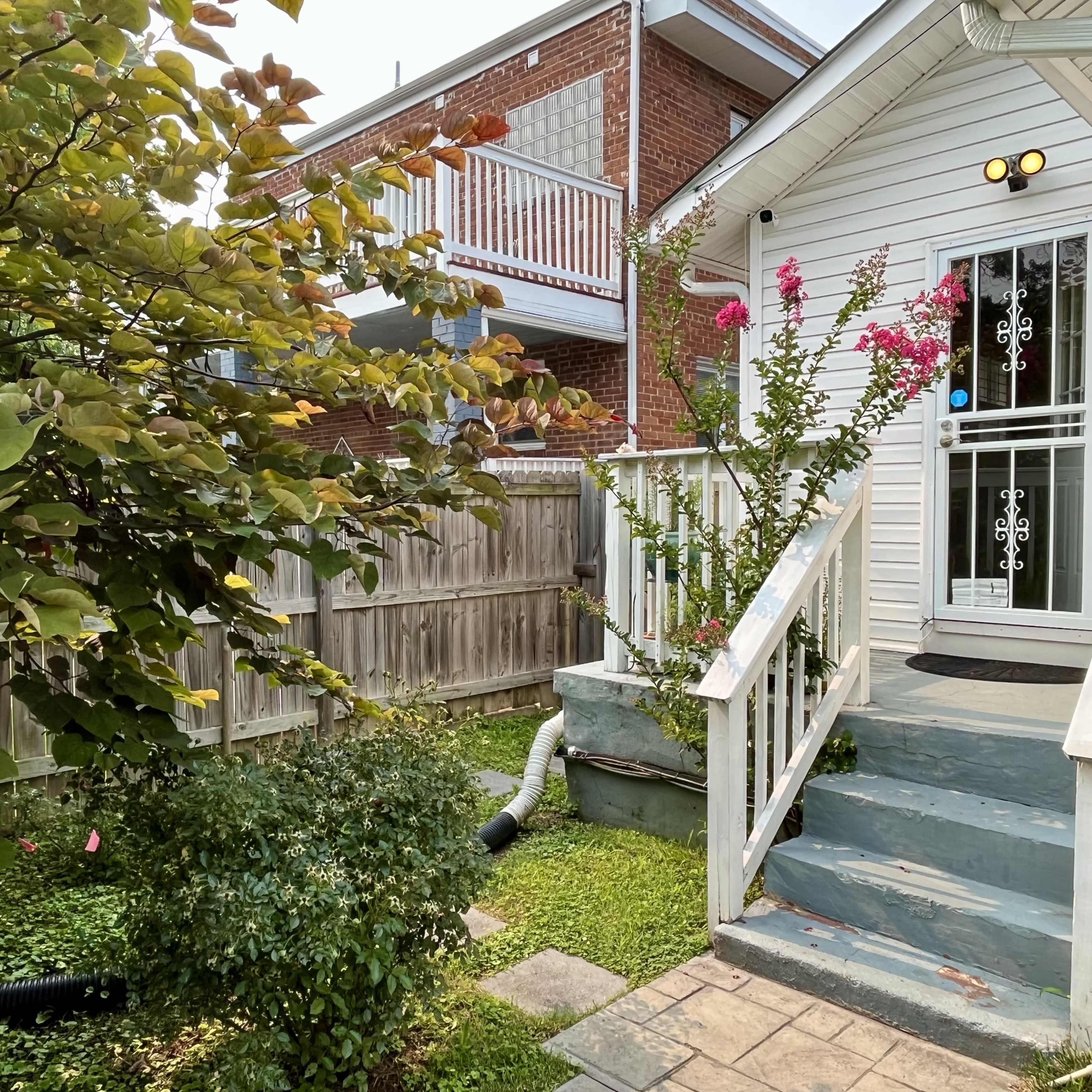 A small, white house with a front porch is surrounded by greenery and a fenced yard, featuring pink flowers and drainage pipes.