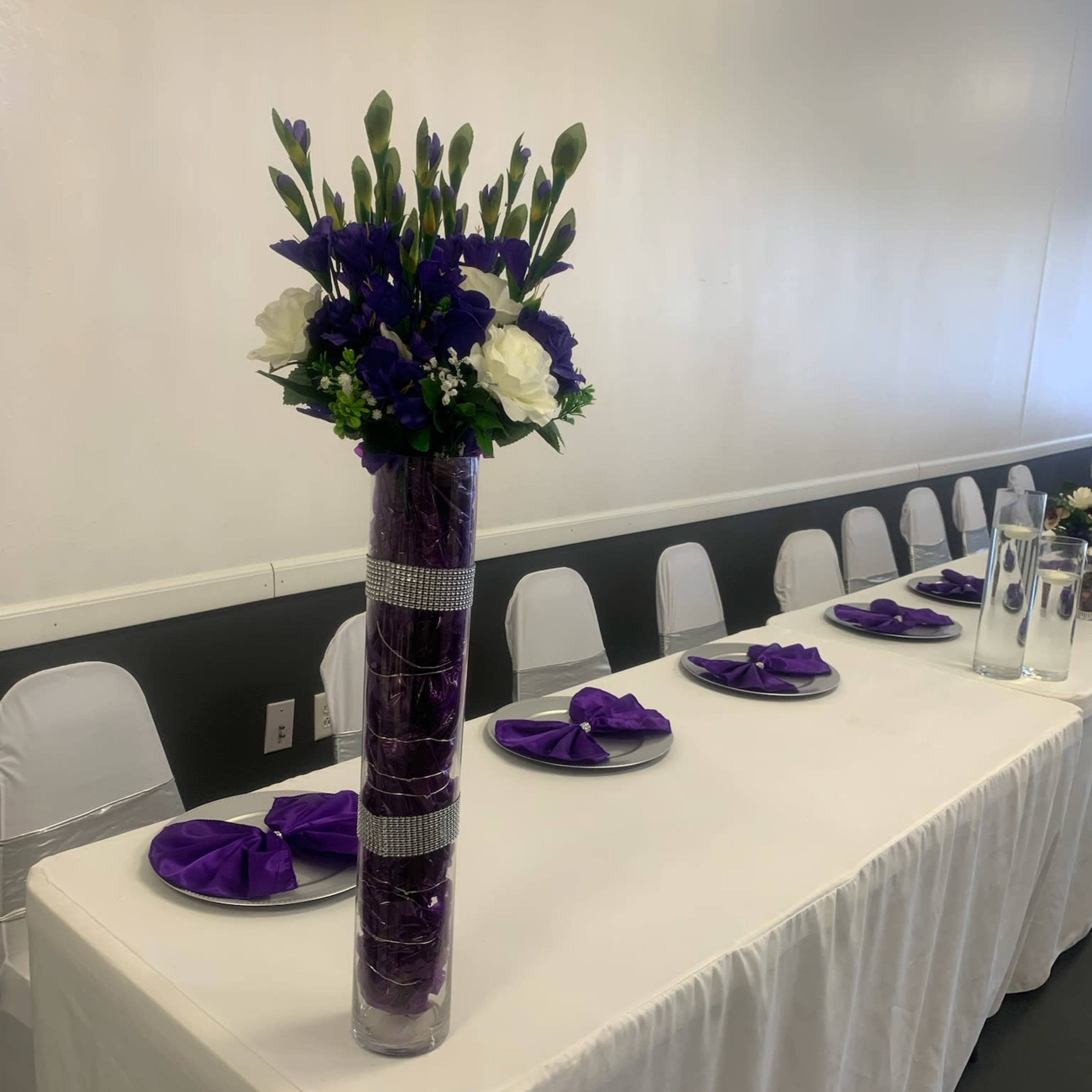A decorated banquet table features a tall floral arrangement and elegantly folded purple napkins on plates.