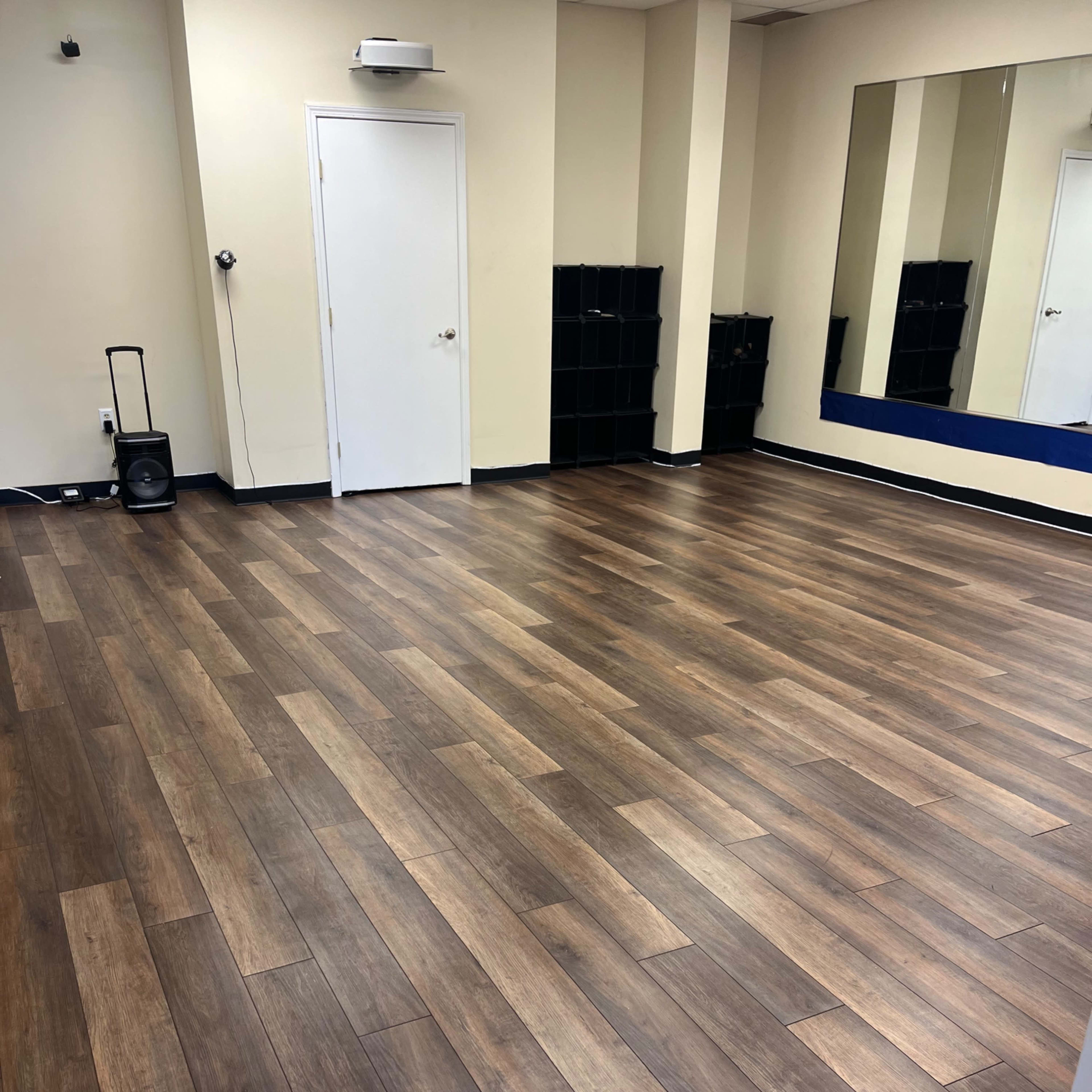 A spacious, empty room with wooden flooring, a mirror on the right wall, and a door in the back.