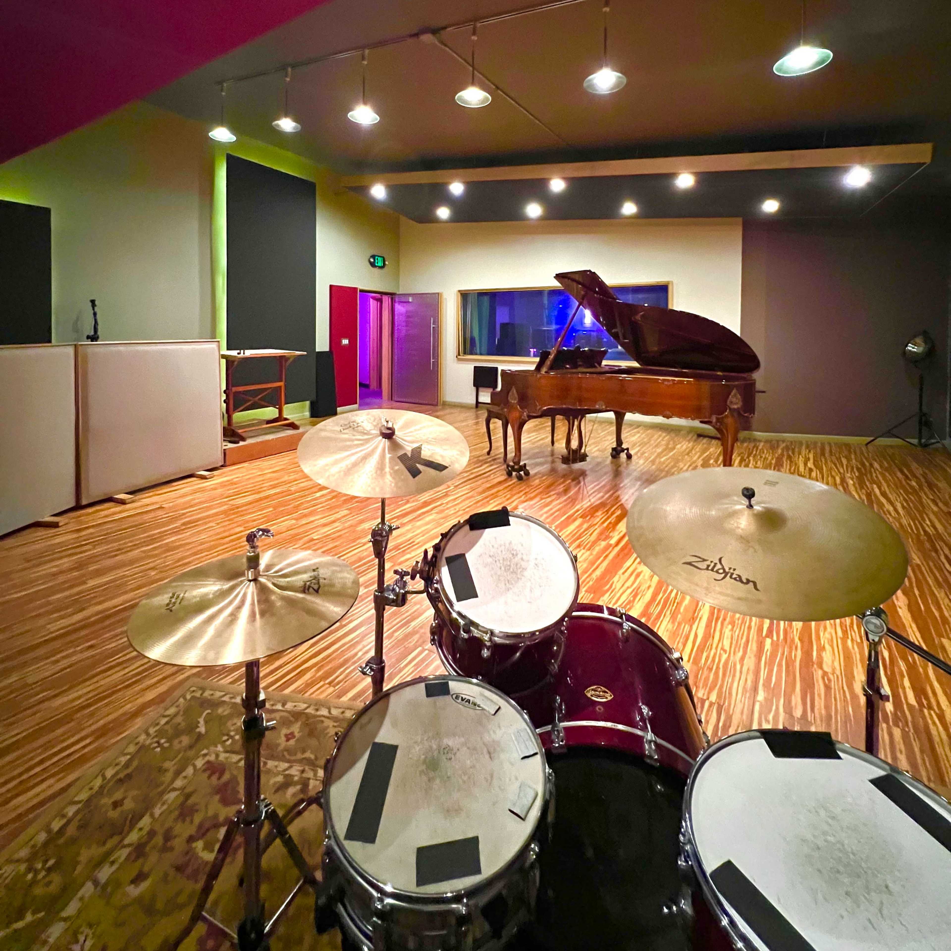 A music recording studio with a drum set in the foreground and a grand piano in the background.