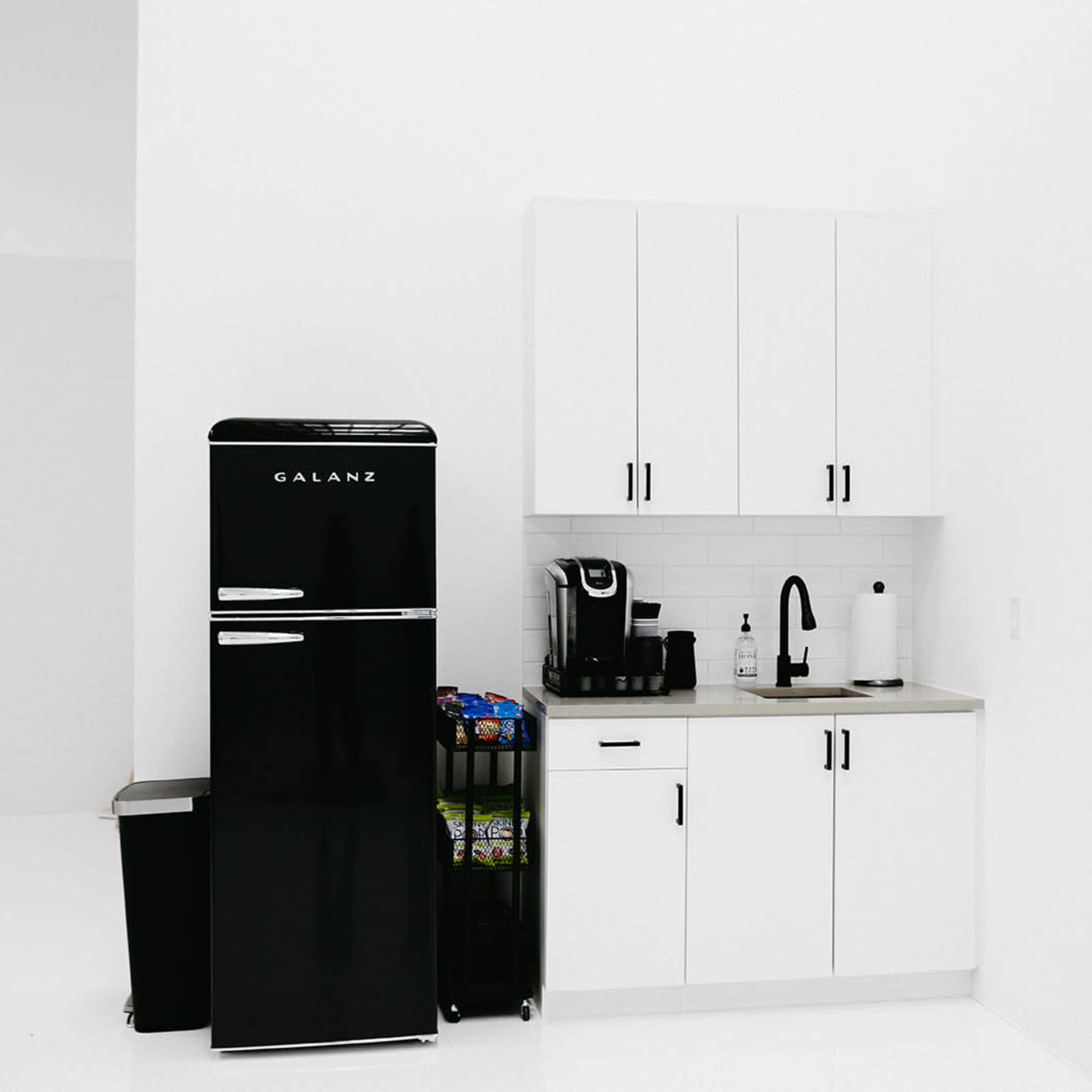 A minimalistic kitchen setup with a black refrigerator, a coffee maker, and white cabinets against a bright backdrop.