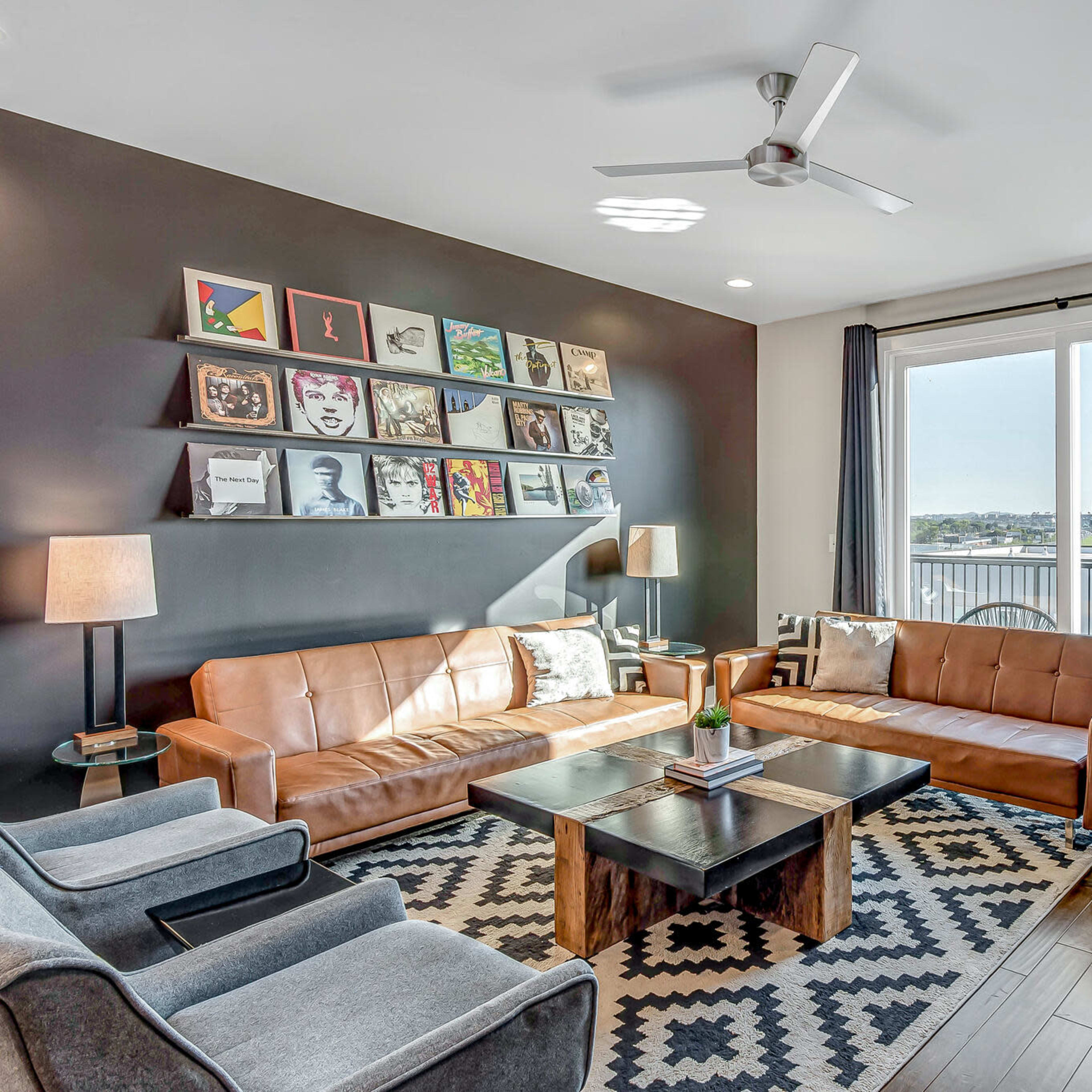 A modern living room features two leather sofas, a coffee table, and a wall adorned with framed artwork, with large windows offering a view of the skyline.